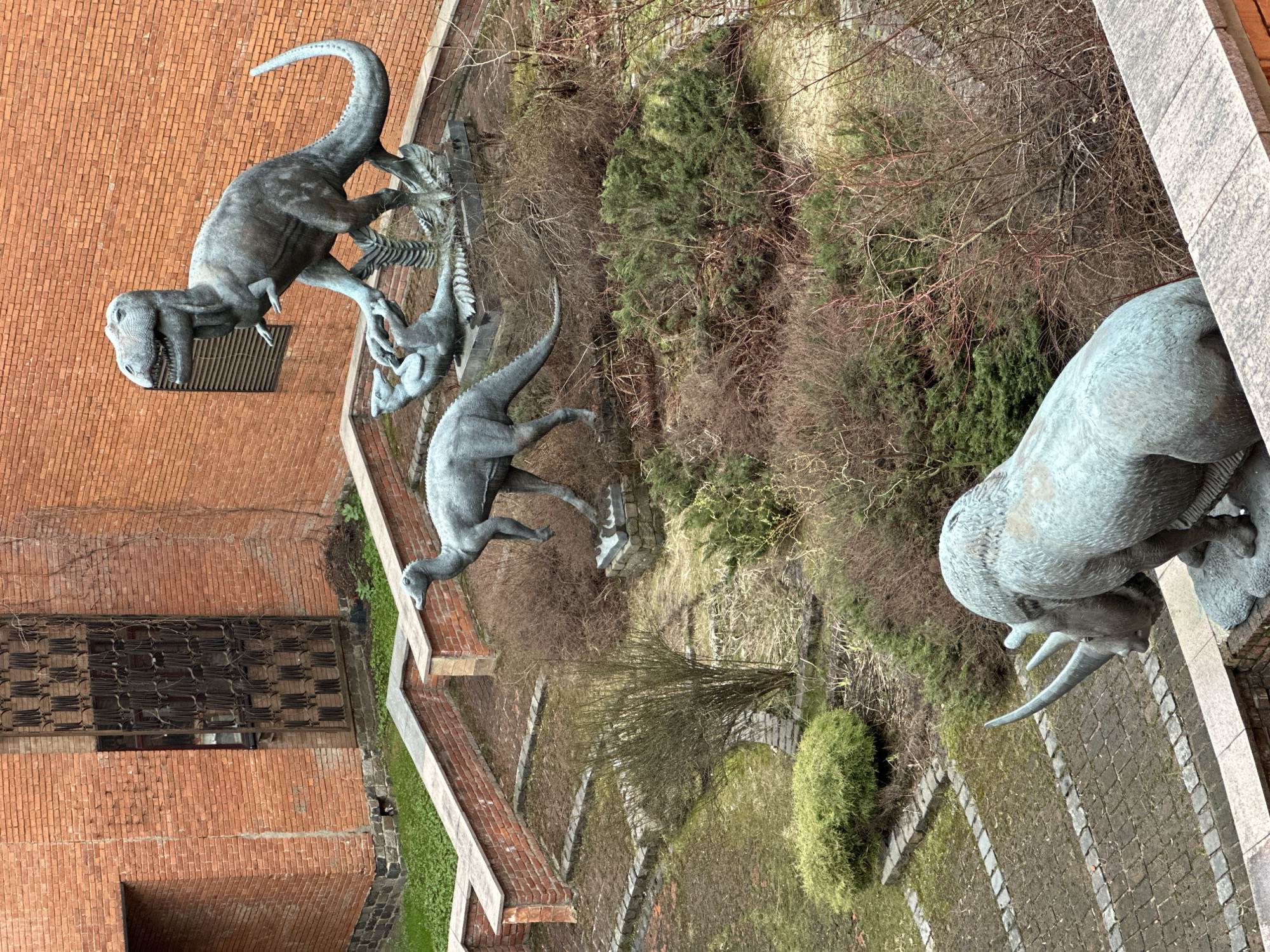 The image depicts the Moscow Paleontological Museum, showcasing bronze dinosaur sculptures arranged in an outdoor setting. The sculptures include a Tyrannosaurus Rex, several smaller dinosaurs, and a mammoth. The background consists of brick buildings and tiered landscaping with greenery and bare trees, reflecting an autumn or early spring atmosphere. The view is taken from above, offering a comprehensive perspective of the layout.