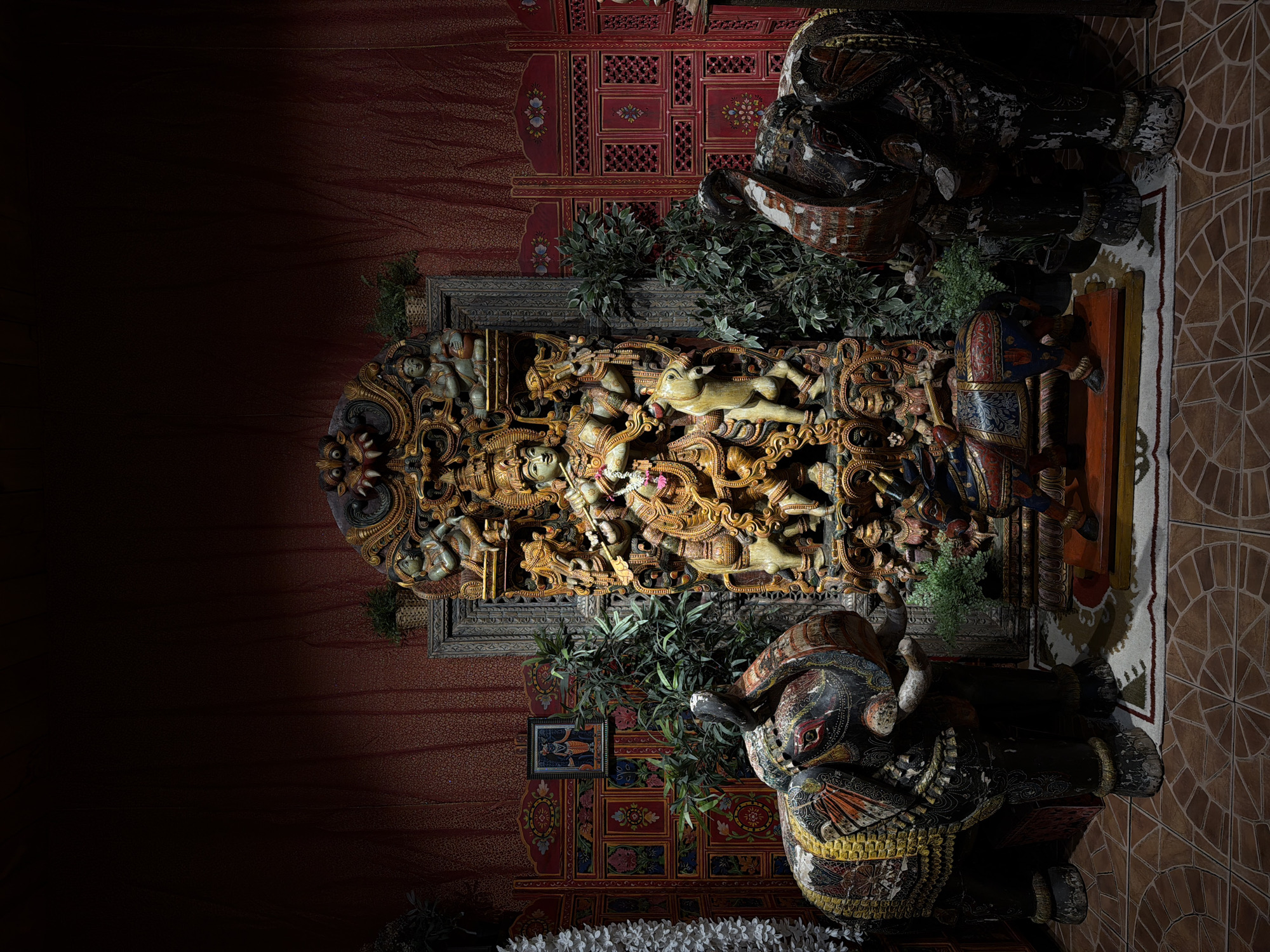 The photograph depicts an indoor shrine with an elaborate, carved wooden deity at its center, possibly representing a Hindu god such as Krishna, adorned with jewelry and playing a flute. Flanking the central deity are large, intricately painted wooden elephant statues, each with decorative coverings. In front of the deity is a smaller carved sculpture of a horse with a rider. Green foliage is strategically placed around the statues, and behind them, a textured red fabric backdrop is visible, along with ornate red screens on either side. A small framed picture hangs on the left screen. The floor is tiled in a circular pattern. The lighting is dramatic, with the central area brightly illuminated and the surroundings in shadow, creating a theatrical atmosphere. The scene is located in the room of the "ISKCON Moscow" temple, which is situated in the city of Moscow, Russia.