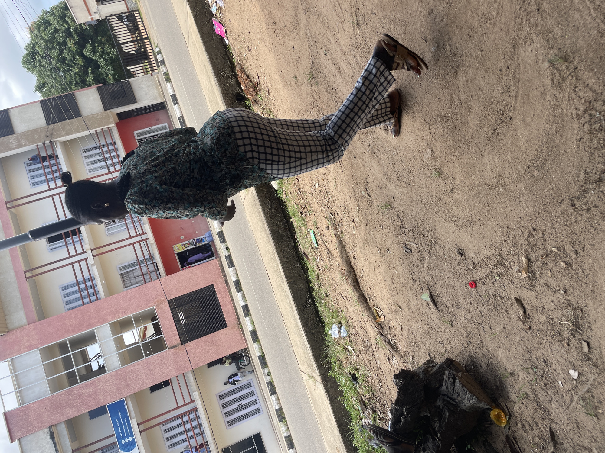 The image depicts a scene on a paved street in Jos, Nigeria, with a pink and white building on the left. A woman wearing a green and black patterned top and checkered pants is walking away from the camera on the right side of the street. She is wearing brown sandals. In the background, several people are visible near the building, and a motorcycle is parked. The street is lined with a concrete gutter and a patch of overgrown grass and dirt. Various pieces of litter are scattered on the ground, including plastic bottles and leaves. A large piece of dark wood is on the left side of the frame in the foreground. The sky is overcast with clouds.