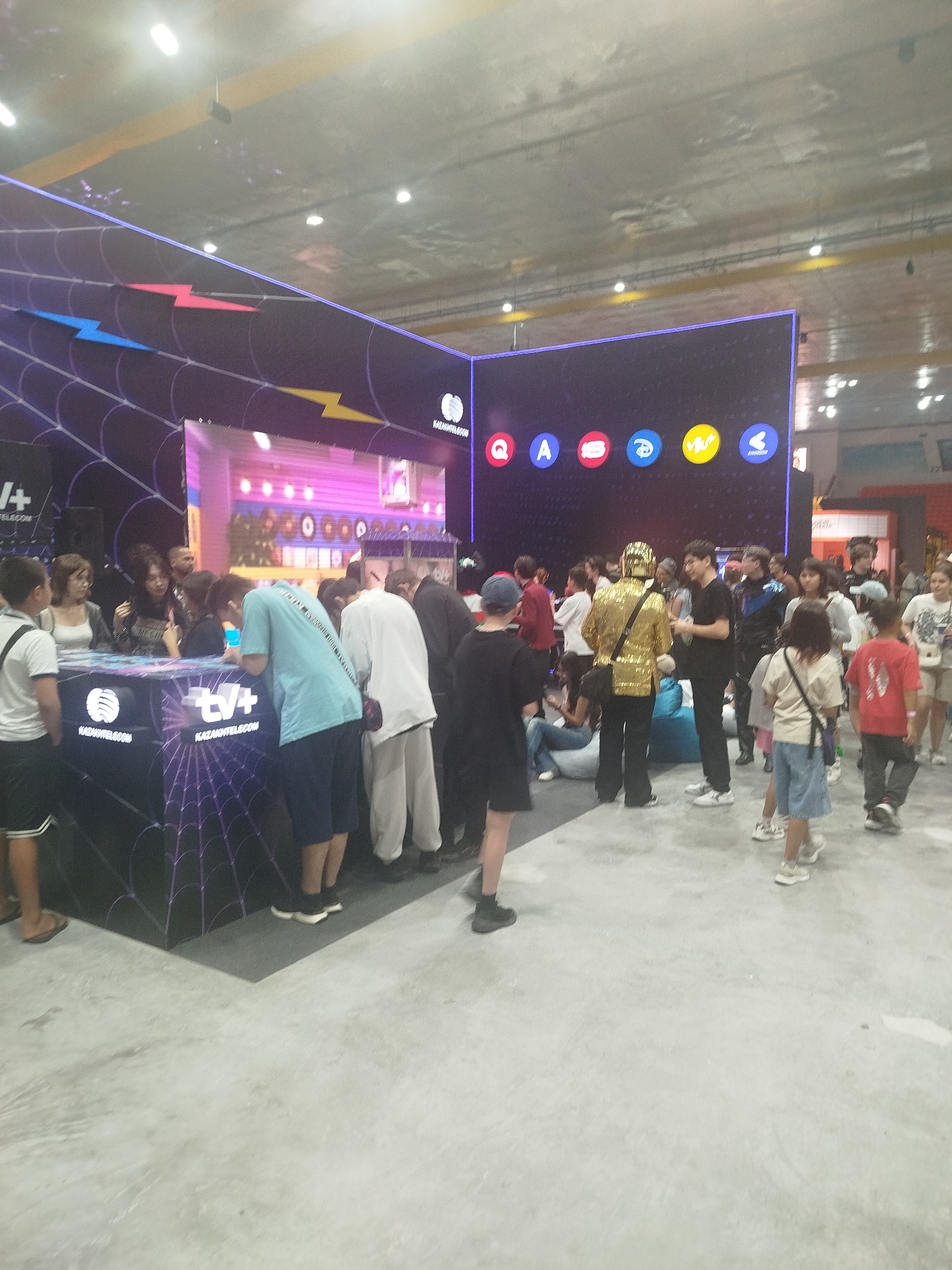 This image captures a lively indoor event, likely a technology or entertainment expo, taking place at the EXPO 2017 complex in Astana, Kazakhstan. The scene is dominated by a large, brightly lit digital display screen showing various colorful icons, including what appear to be social media or service logos. In front of the screen, a group of people are gathered, some interacting with staff or displays. A prominent booth features the "TV+" and "KAZAKHTELeCOM" branding, adorned with a spiderweb-like design.
Several individuals are visible in the foreground and midground, dressed in casual attire. Among them, a person in a striking gold, sequined jacket stands out, drawing attention. A young boy in a white t-shirt and black shorts is on the left, facing away from the camera. To his right, a group of people are clustered around the "TV+" booth. Further back, more attendees mill about, some looking at the large screen, others engaged in conversation or observing the activities. The environment suggests a convention center or exhibition hall, with a high ceiling and industrial lighting. The overall atmosphere is one of engagement and public interaction with modern technology and entertainment.