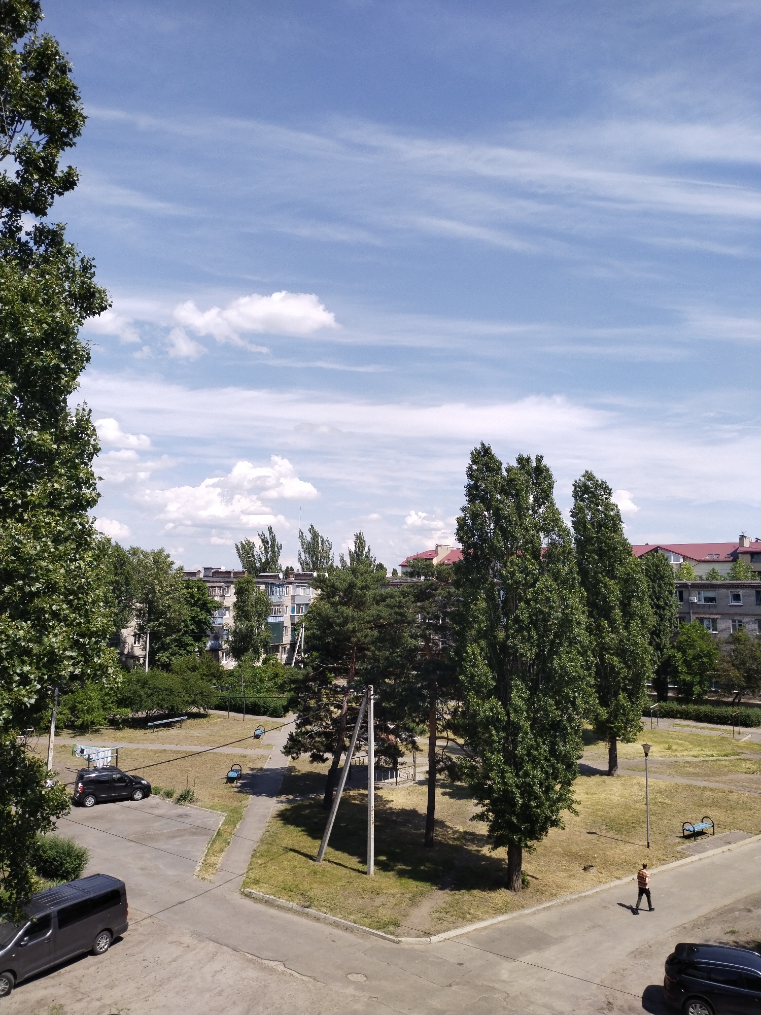 This image captures a high-angle view of a residential area in Horishni Plavni, Ukraine, under a clear blue sky with scattered stratocumulus and cirrus clouds. A large, leafy green tree partially obstructs the left side of the frame in the foreground.
Below the tree, an asphalt parking area is visible, with a dark grey minivan parked facing right and a black compact SUV/hatchback parked facing left. A concrete path network traverses a grassy, partially dry park area, where several light blue metal benches are situated. A prominent utility pole with multiple overhead wires stands upright in the center-left midground. Several tall, slender deciduous trees with dense green foliage and a few coniferous trees are scattered throughout the park area and further into the background.
On the right side, a person wearing a horizontally striped shirt (orange, white, and dark) and dark trousers is seen walking on an asphalt road away from the viewer. A grey street light pole stands to the left of the pedestrian. A dark SUV is partially visible, parked on the far right edge of the frame.
In the background, beyond the trees, several multi-story residential buildings with various roof types (some red-tiled, some flat) and visible windows/balconies are discernible. The overall scene is brightly lit by daylight, casting distinct shadows.