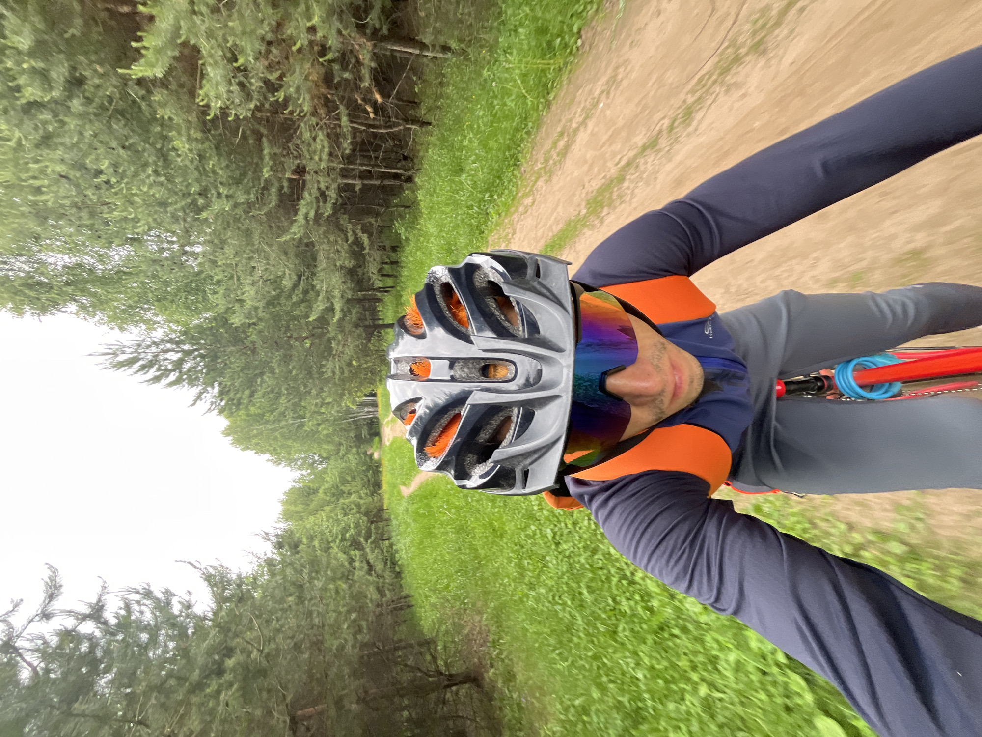 In Chaikovsky, Russia, a person on a bicycle rides along a dirt path through a forest. The individual is wearing a dark long-sleeved shirt, grey pants, an orange and grey helmet, and reflective sunglasses. The path is bordered by lush green grass and tall pine trees. The sky is overcast.