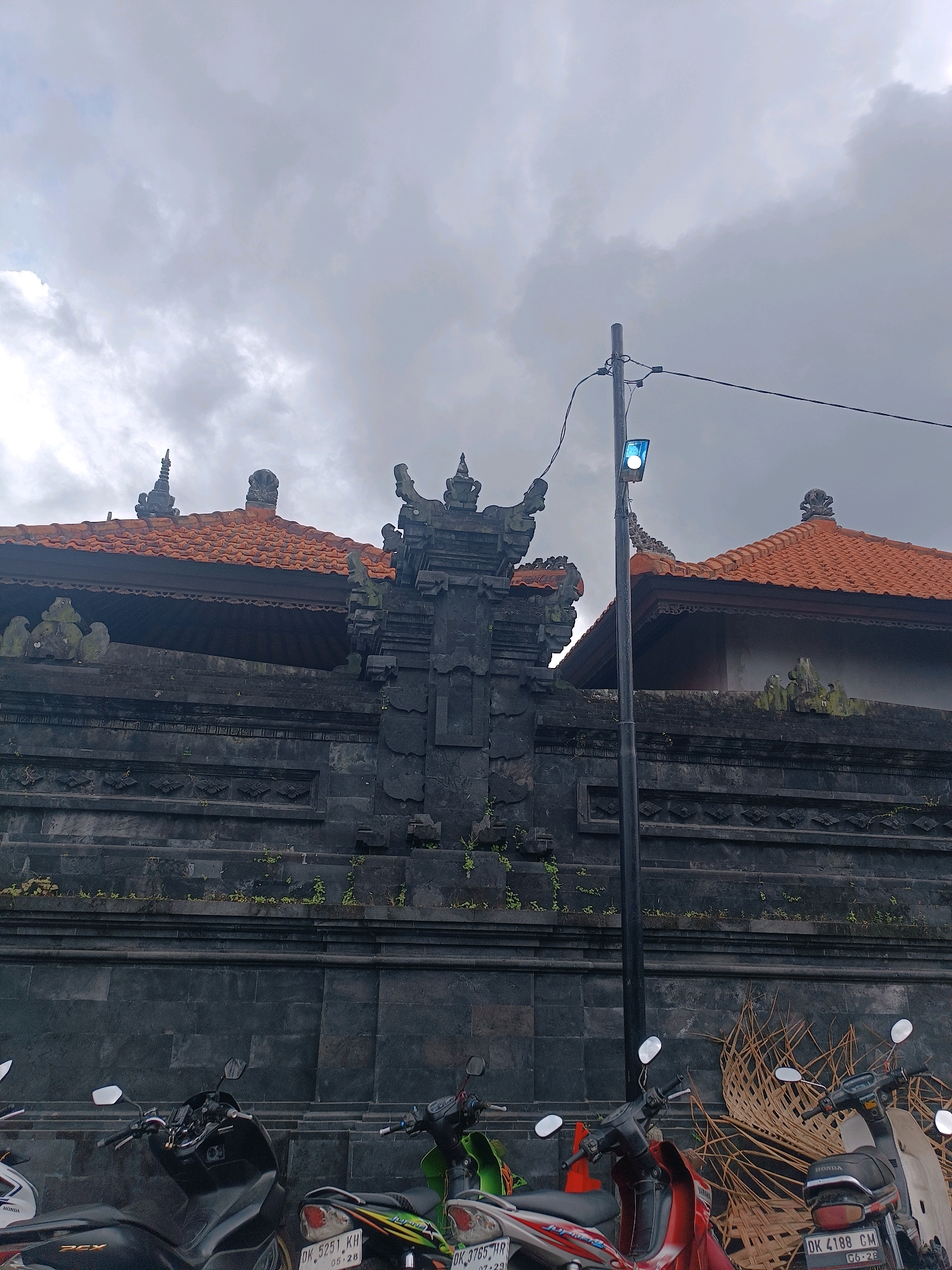 In Selemadeg, Indonesia, a gray, cloudy sky looms over a dark, stone wall with ornate carvings. The wall has an orange-tiled roof on top. A street light with a bright blue bulb stands to the right of the wall, with wires extending from it. Several parked motorcycles are visible in front of the wall. The scene appears to be an outdoor, public space.