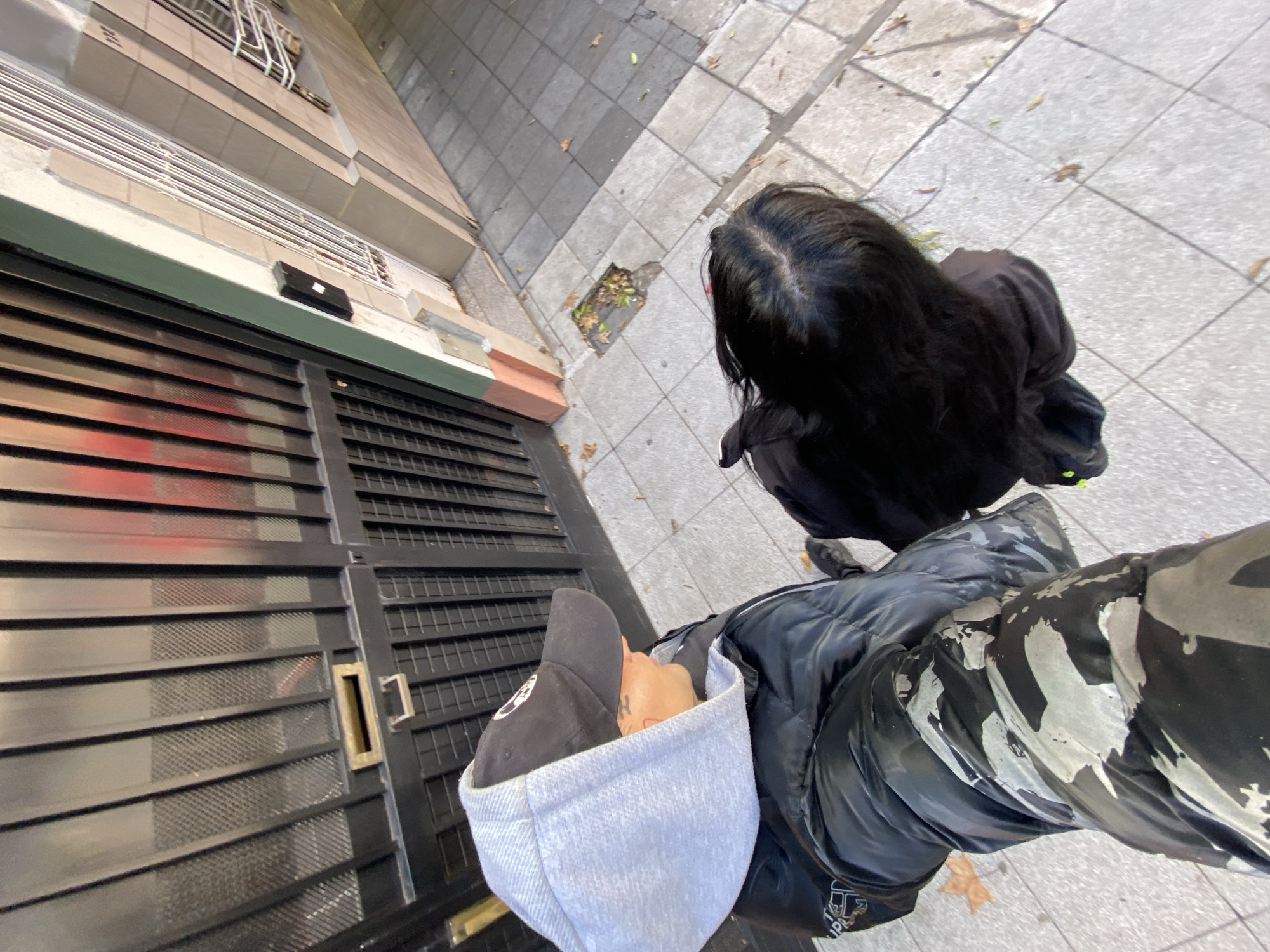 The image depicts a street scene in Buenos Aires, Argentina. The perspective is from a high angle, possibly taken from a balcony or upper floor. The foreground features two individuals. One person wears a black baseball cap with a light grey hoodie, a black jacket, and camouflage pants. The second person, positioned slightly ahead, has long, dark hair and is wearing a black jacket.
The background shows a building with a black metal gate. The gate has horizontal slats with a decorative grille behind them. Adjacent to the gate is a section of the building's façade in beige, light orange, and brown. The sidewalk is paved with grey tiles and has some fallen leaves. A small, empty square space can be seen in the pavement near the building.