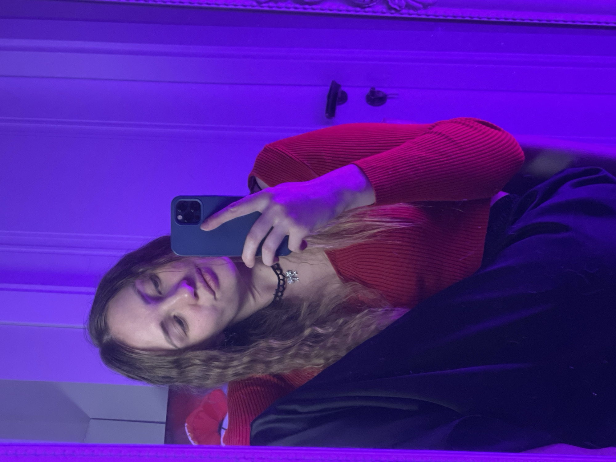 A young person takes a selfie with a blue mobile phone. They are wearing a red, long-sleeved, ribbed shirt and black pants. They also wear a black choker with a silver snowflake charm. The background is a white wall with moldings and a purple light illuminating the scene. The location cannot be determined from the image.