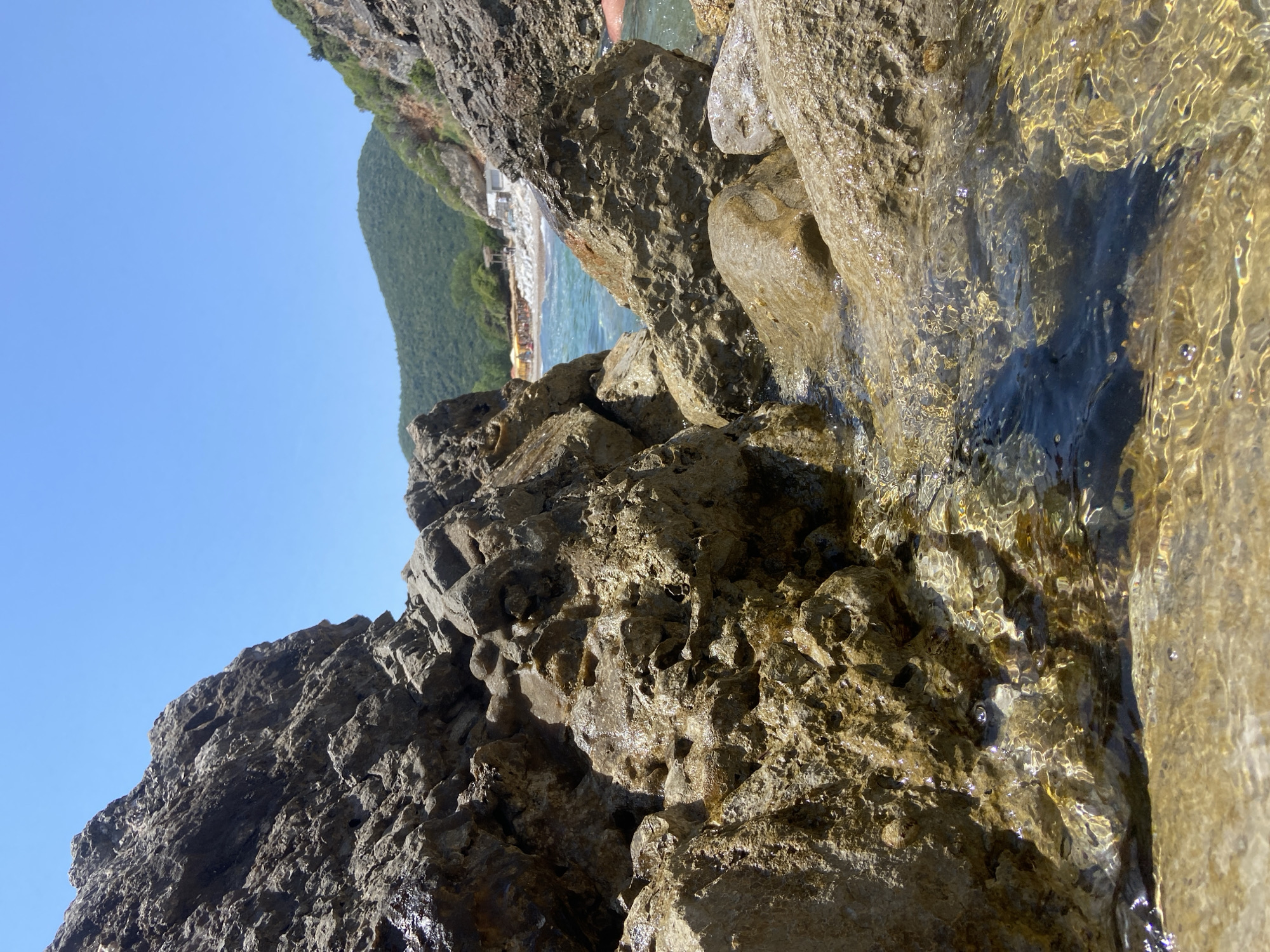 The image depicts a rocky coastline with clear, shallow water flowing over the rocks. In the background, a beach with people and buildings is visible, backed by a green, forested hill. The sky is clear and blue. This scene appears to be located near the city of Prijevor, Montenegro.