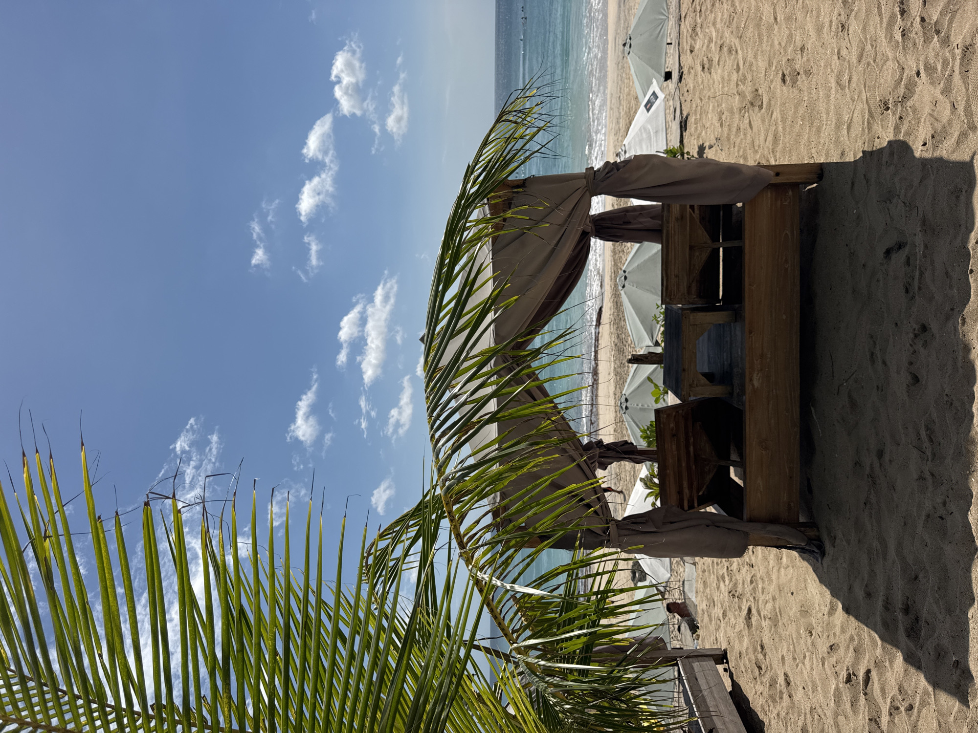 The image depicts a beach scene in Kuta Selatan, Indonesia. A wooden cabana with beige curtains sits on the sand. Beyond the cabana are white beach umbrellas and the ocean. A palm frond is visible in the foreground, partially obscuring the cabana. The sky is blue with scattered clouds. The sand shows footprints and shadows. The lighting suggests a sunny day.
