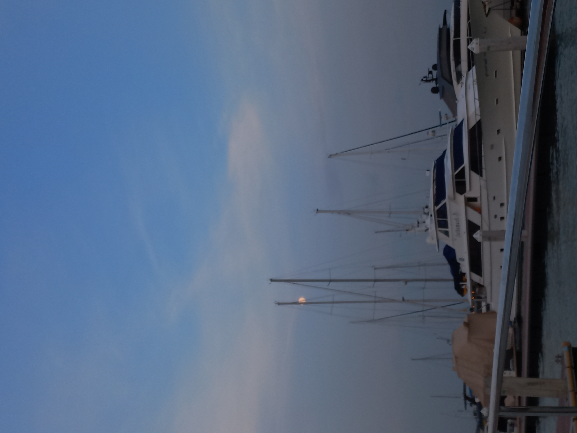 The image displays a serene twilight scene at a marina, likely in Na Chom Thian, Thailand. The upper and left portions of the frame are dominated by a vast expanse of sky, transitioning from a clear, deep blue on the far left to a lighter, hazy blue-gray towards the right and bottom, with faint, wispy clouds softly accentuating the horizon.
In the lower and right parts of the image, several boats are moored alongside a pier or jetty. These include both white motor yachts and sailboats, with their intricate masts and rigging prominently silhouetted against the sky. A notable detail is a hazy, glowing full or near-full moon, visible positioned between two of the sailboat masts, indicating either early morning dawn or late evening dusk.
The largest vessel, a white motor yacht docked nearest the viewer on the right, features sleek lines. Faint white lettering on its hull spells out "MY SYMPHONY." To the immediate right, a section of the dock structure, including a metal railing and sturdy wooden posts, is visible, with dark, calm water reflecting the subdued light.
There are no people visible in the frame, contributing to the quiet and tranquil atmosphere. The scene suggests peaceful weather conditions, with the soft lighting creating a calm and contemplative mood over the marina.
