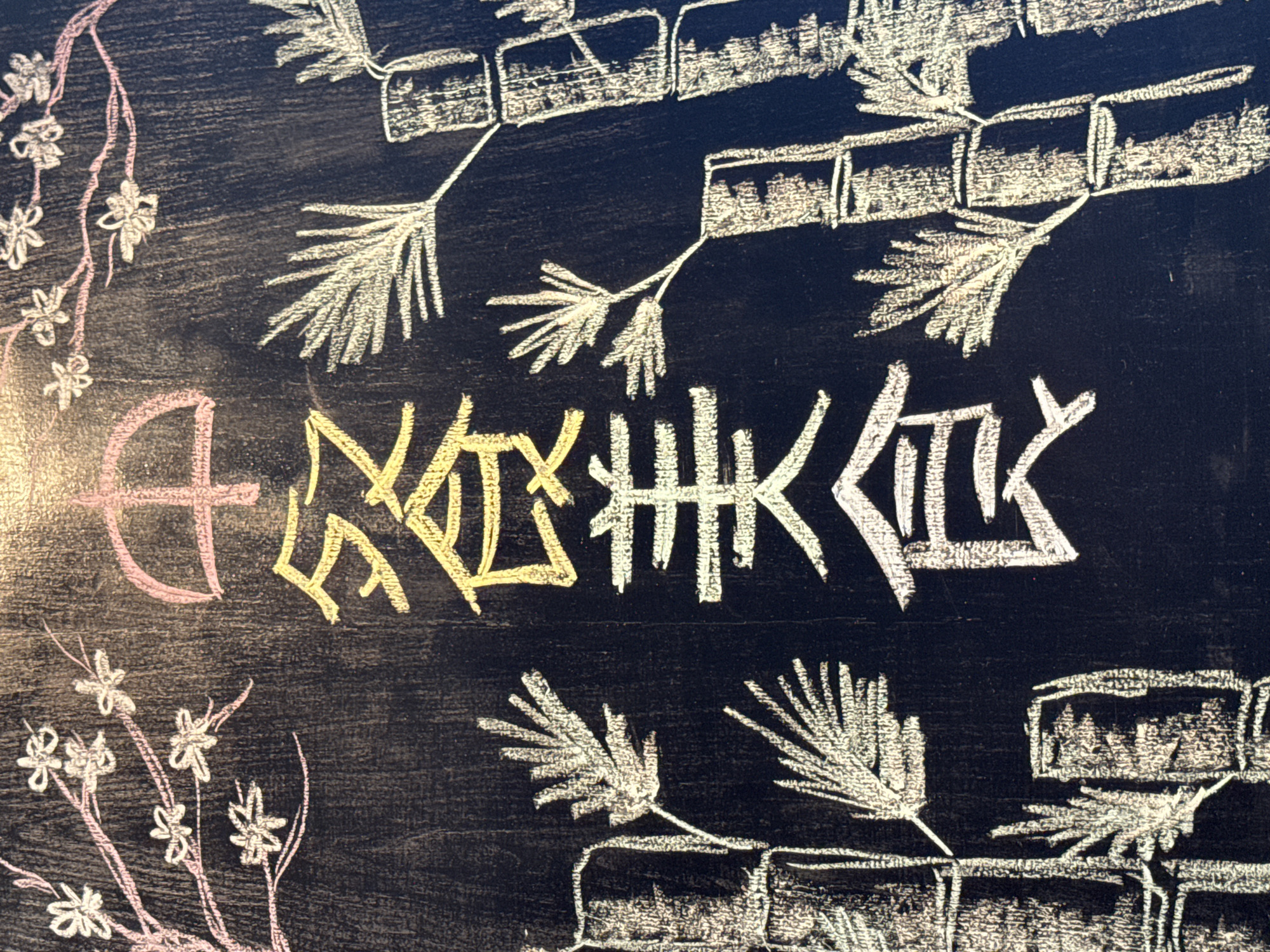 The image shows a blackboard with chalk drawings of Chinese characters and bamboo stalks. The characters appear to be "餐食" (cān shí), which translates to "food" or "meal." The bamboo is drawn in white chalk with segmentation lines indicating the different nodes. To the left of the characters, there are delicate pink chalk drawings of cherry blossoms on a branch. The blackboard surface has a rough texture with visible vertical grain lines. The lighting suggests an overhead light source, casting a slight glow at the top center. The overall composition is artistic, likely intended as decoration or signage. The provided context states the image is located in Kuta Selatan, Indonesia.