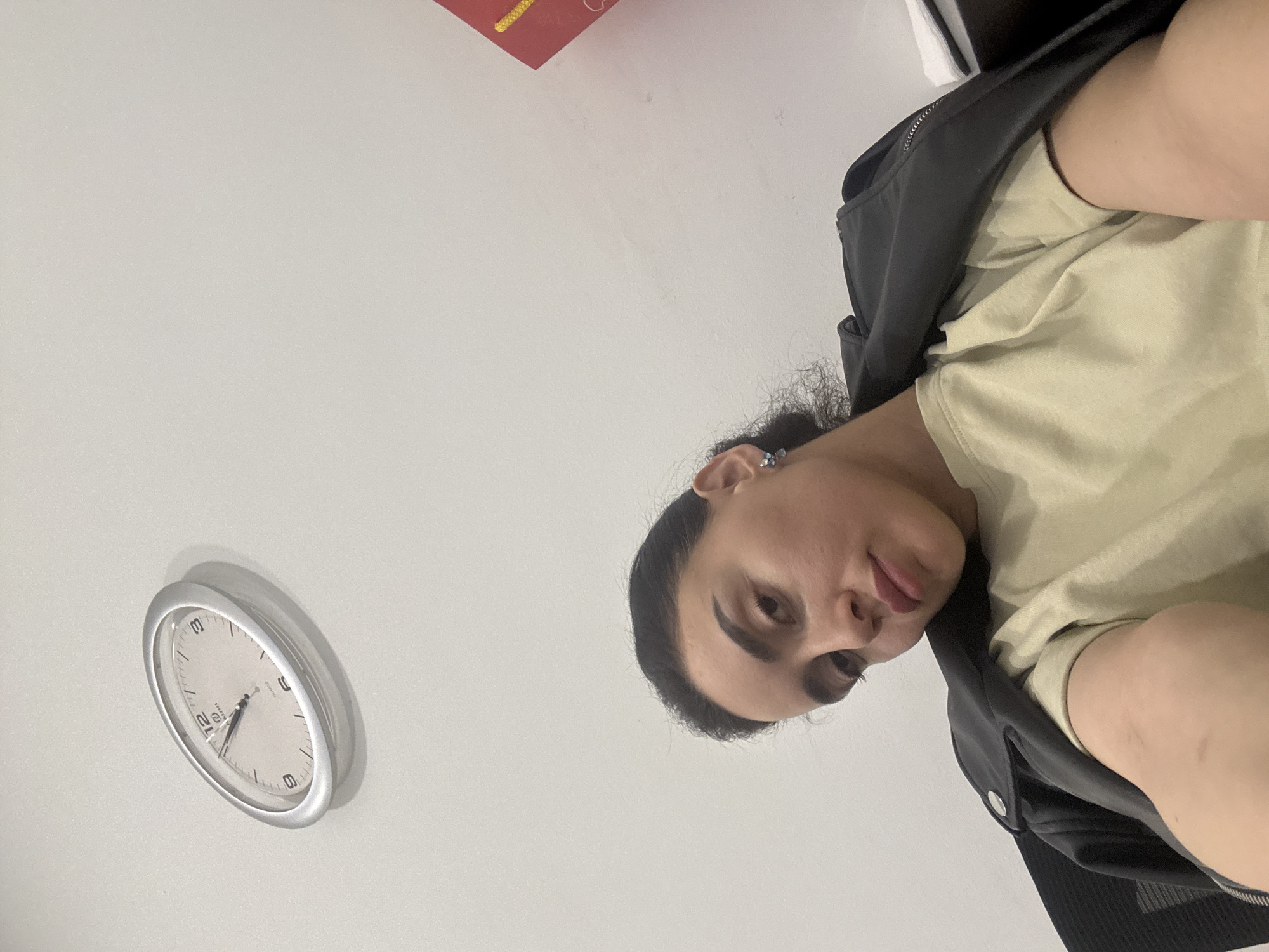 A selfie taken by a person with dark hair wearing a light-colored shirt and a black jacket. The person is positioned in the upper right of the frame. A clock on a white wall is visible in the lower left corner, showing approximately 7:25. Part of a red object is present in the upper portion of the image. The location is not discernible from the image.