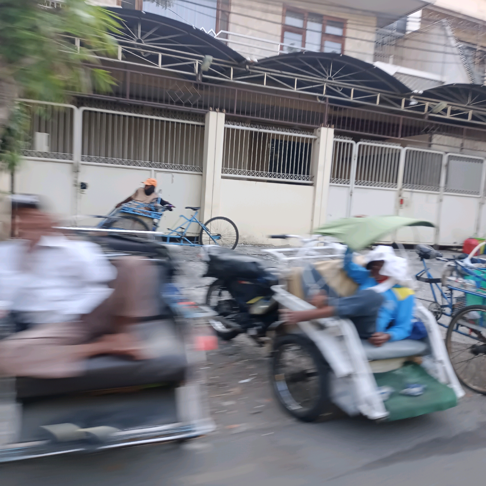 A street scene in Surabaya, Indonesia, shows blurred figures and vehicles in motion. In the foreground, a person in a white shirt is seated in a three-wheeled vehicle, likely a becak or motorized rickshaw. To the right, another becak is visible, with a person in a blue jacket and yellow trim seated under a green canopy. A third becak with a rider wearing an orange cap is further back. In the background, a building with a metal awning and barred windows lines the street, with a white fence in front. The ground is paved and appears wet or dusty, with scattered debris. The overall impression is one of a busy street with ongoing traffic.
