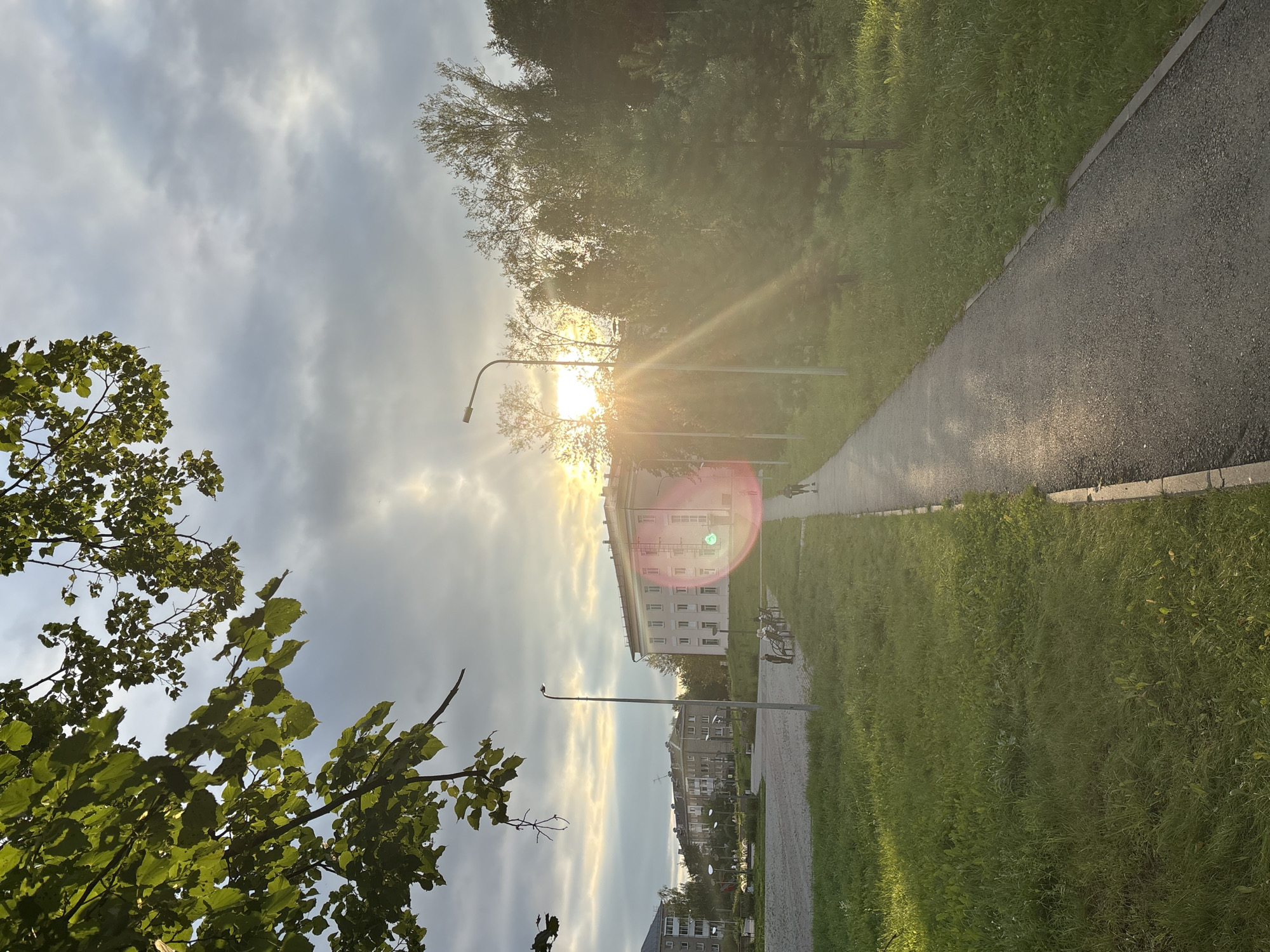 The image depicts an outdoor scene in the city of Krasnokamsk, Russia, likely in the late afternoon or early evening due to the low sun angle and lens flare. In the foreground, a paved pathway runs diagonally from the lower right to the mid-left of the frame, bordered by a strip of grass on the right and a wider expanse of grass on the left. Tall green grass fills the left side of the pathway.
Several buildings are visible in the background. To the left, there are multi-story residential buildings with a similar architectural style, characteristic of Soviet-era construction. In the mid-ground, a longer, lighter-colored building with multiple windows and balconies is prominent. To the right of this building, a dense line of trees, including evergreen and deciduous varieties, obscures some of the buildings behind them.
A single street light stands near the center of the image, just to the left of the pathway. The sun is setting behind the trees on the right, casting a strong lens flare across the image, particularly in the mid-right section. The sky is overcast with dramatic clouds, but the sun is breaking through in the upper center and mid-right, creating bright rays.
Several individuals are present. On the pathway, a lone person is walking away from the camera towards the mid-ground. Further back, near the lighter-colored building, a group of people are seated on benches. In the far left, more figures are visible near the residential buildings. The overall impression is a residential area with a park or green space, suggesting a common urban landscape in Krasnokamsk.