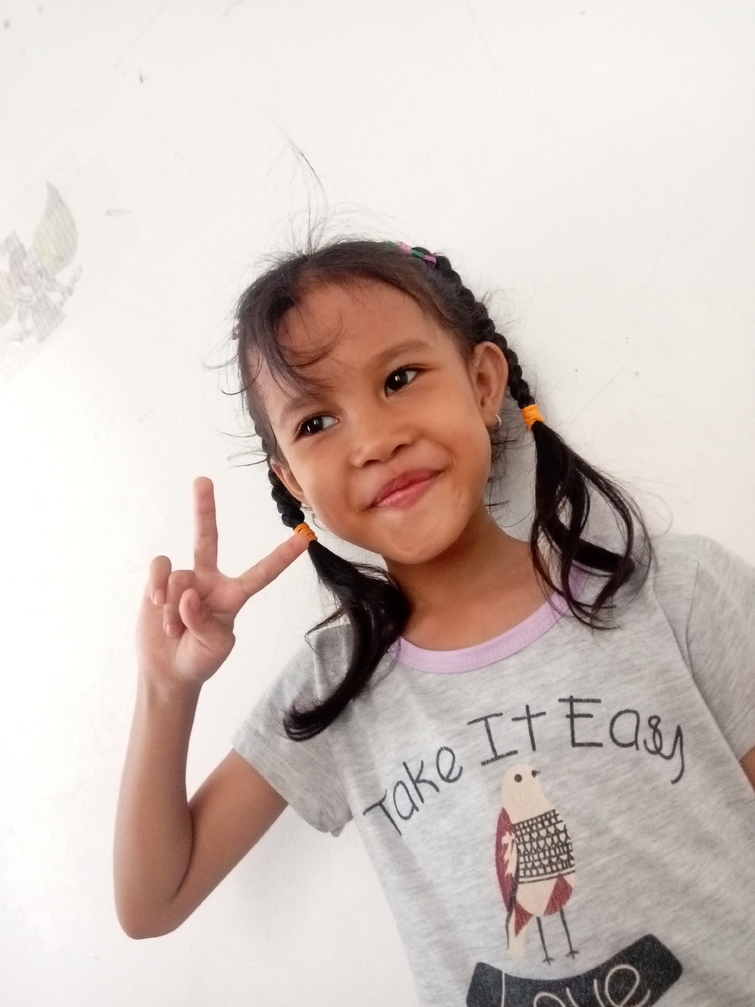 A young girl with pigtails is posing for a photo against a white wall. She is wearing a grey t-shirt with a drawing of a bird and the words "Take It Easy". She is holding up two fingers in a peace sign. The image appears to be taken indoors, possibly in a home, given the plain white wall and the simple setting. There is a faint drawing on the wall behind her, which may be an emblem or artwork. It is not possible to determine the specific location within Batam, Indonesia from the image alone, as the image lacks any distinguishing landmarks or contextual clues that would pinpoint a particular area or establishment in the city. The lighting suggests it might be daytime, possibly near a window or with artificial lighting.