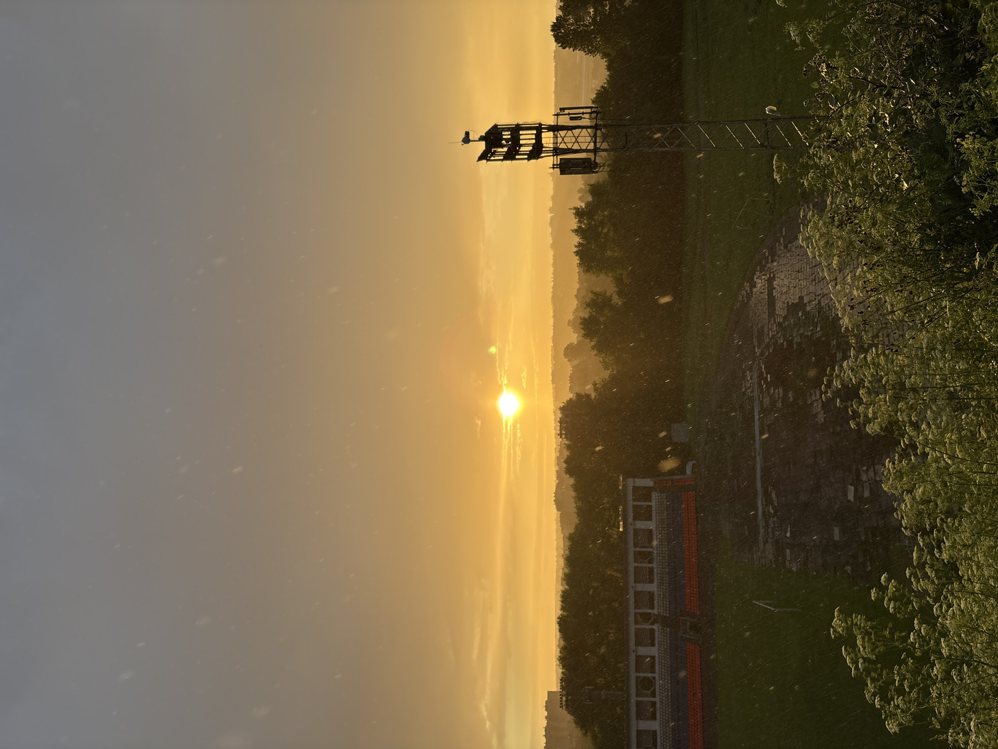 The image depicts an outdoor scene at sunset, likely in the city of Sof'ino, Russia, characterized by heavy rainfall. The sky is dominated by a brilliant orange sunset, with the sun positioned slightly off-center to the right, illuminating the horizon with a warm glow. Wispy clouds are visible, tinged with the colors of the sunset. Small white specks, representing raindrops, are scattered throughout the frame, indicating the ongoing precipitation.
In the foreground, lush green vegetation, including flowering plants and bushes, partially obscures the lower portion of the image. Beyond the vegetation, a sports field is visible. To the left of the field are rows of stadium seating, with a mix of orange and blue-colored seats. Part of the field appears to be paved or a darker material, possibly a track or pathway, with areas of standing water reflecting the light.
Further in the background, a dense line of trees creates a visual barrier, behind which distant buildings and the cityscape of Sof'ino are discernible. To the right of the field, a tall, metallic communication tower stands prominently. The tower is equipped with multiple antennas and what appears to be a small structure or platform at its apex. The overall atmosphere is dramatic and moody, a combination of the beautiful sunset and the persistent rain. No people are visible in the image.