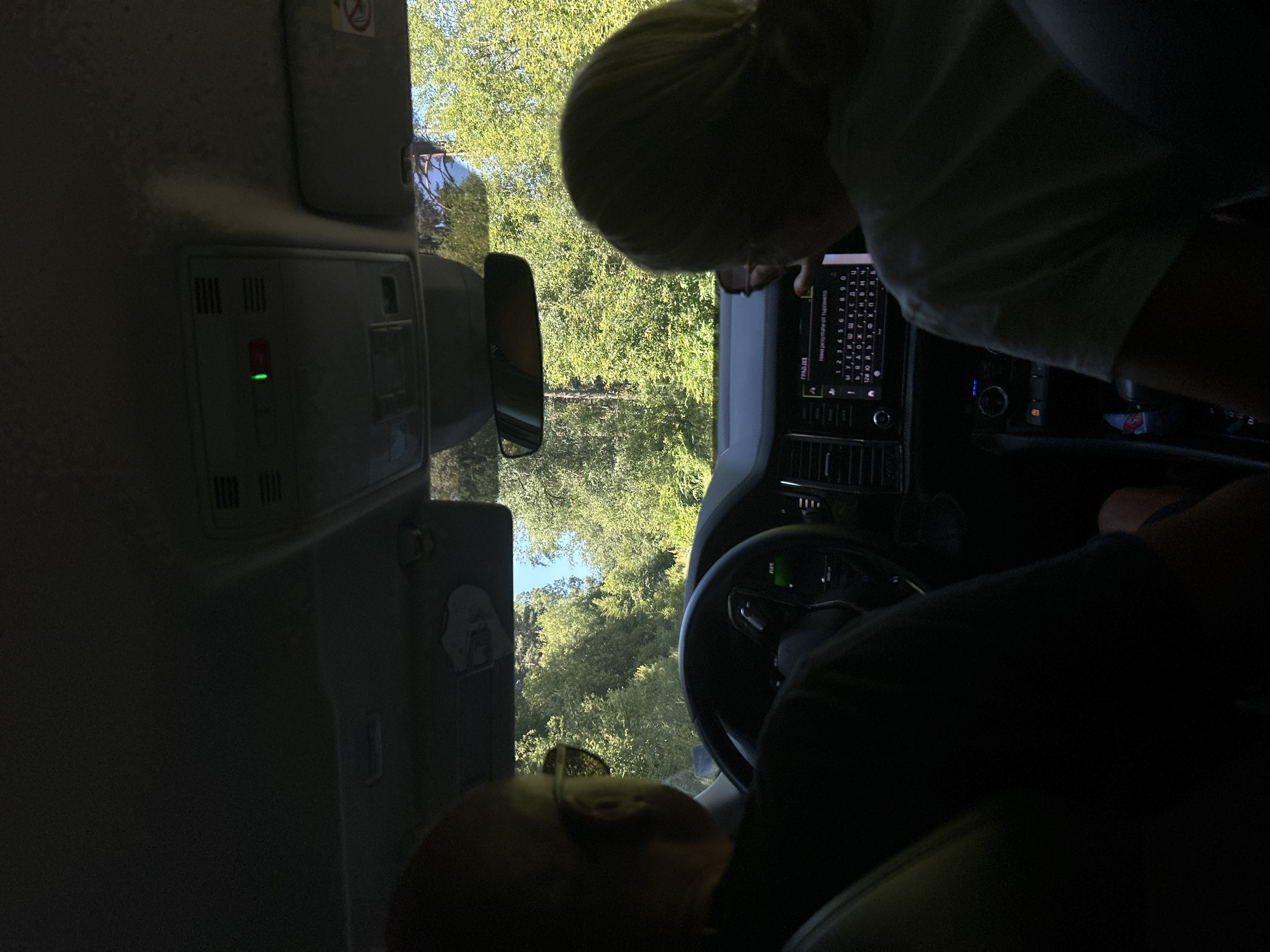 The image shows the interior of a car, viewed from the back seats looking forward. Two individuals are visible in the front seats, a driver and a passenger. The driver, on the left, is wearing sunglasses and is partially obscured. The passenger, on the right, has blonde hair and is looking at the car's infotainment system, which displays a keyboard interface.
The car appears to be in motion, as indicated by the blurred greenery and trees visible through the windshield, suggesting a road surrounded by nature. The sunlight is strong, casting shadows inside the car. The car's dashboard and roof console are visible. A green indicator light is illuminated on the roof console, likely for a safety feature.
The location is specified as the city of Batak, Bulgaria. This suggests the car is traveling on a road in or near this city, likely in a rural or scenic area given the dense foliage. The overall scene depicts a car journey through a verdant environment.
