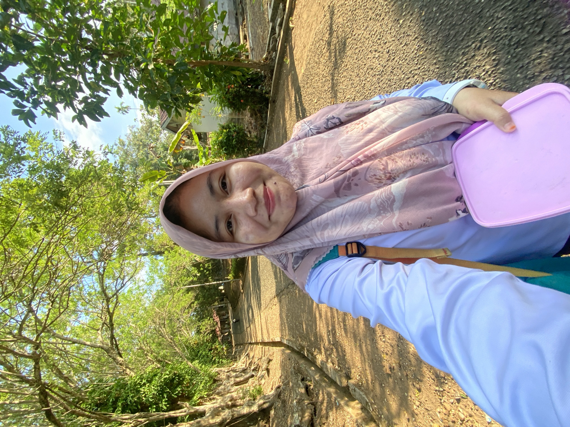 In a selfie taken outdoors, a young woman wearing a light pink hijab and a white long-sleeved shirt smiles at the camera. She is holding a rectangular pink container in her right hand. A backpack with orange straps is visible on her back. The background shows a paved pathway lined with trees on both sides, suggesting a park or a tree-lined street. In the distance, there are buildings and more vegetation, indicative of a semi-urban or rural setting, possibly in Banjarbaru, Indonesia, given the lush tropical foliage and architectural style of the buildings. The lighting suggests it is daytime, with sunlight filtering through the trees.