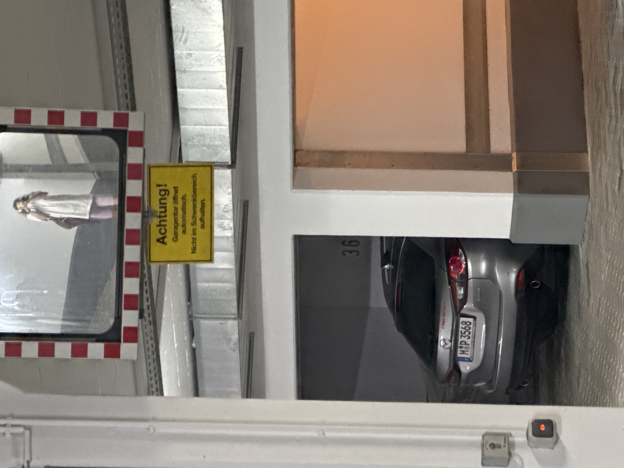 The image depicts an indoor scene, likely a parking garage in Höhenkirchen-Siegertsbrunn, Germany, given the license plate. The orientation of the image is vertical, making it appear as if it was rotated 90 degrees counter-clockwise.
The main subject is a silver car, a Mazda, parked in a designated spot. The car's rear is visible, displaying a license plate with the characters "M&P 3568". The parking spot is marked with the number "36". To the left of the car, a person is reflected in a large mirror or glass panel. This person appears to be wearing a light-colored top and pink leggings, and is facing away from the viewer.
A yellow warning sign is prominently displayed on a metal structure to the left of the car. The text on the sign reads "Achtung! Garagentor öffnet automatisch. Nicht im Schwenkbereich aufhalten." which translates to "Attention! Garage door opens automatically. Do not stop in the swing area." This suggests that the garage door is automated and poses a potential hazard.
The setting is typical of a multi-story parking facility, with concrete walls and metal structural elements visible. The lighting appears to be artificial, indicating it is likely daytime or evening. There are no obvious indications of weather conditions. The overall atmosphere is utilitarian and functional, typical of a parking environment.
