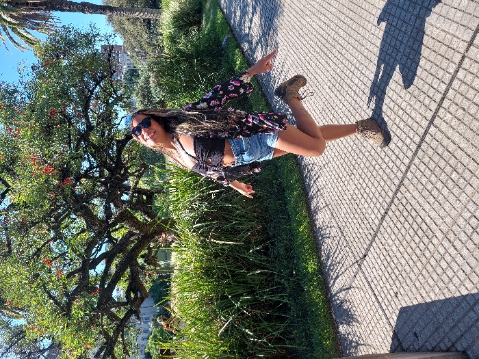 A young woman poses outdoors, wearing sunglasses, a black top, denim shorts, and a floral shirt. She stands on a brick-paved path, with a lush green park and a large tree on her left. The sun casts shadows on the path, suggesting it is a bright day. Buildings are visible in the background. The composition captures a moment of leisure and enjoyment in a natural setting.
