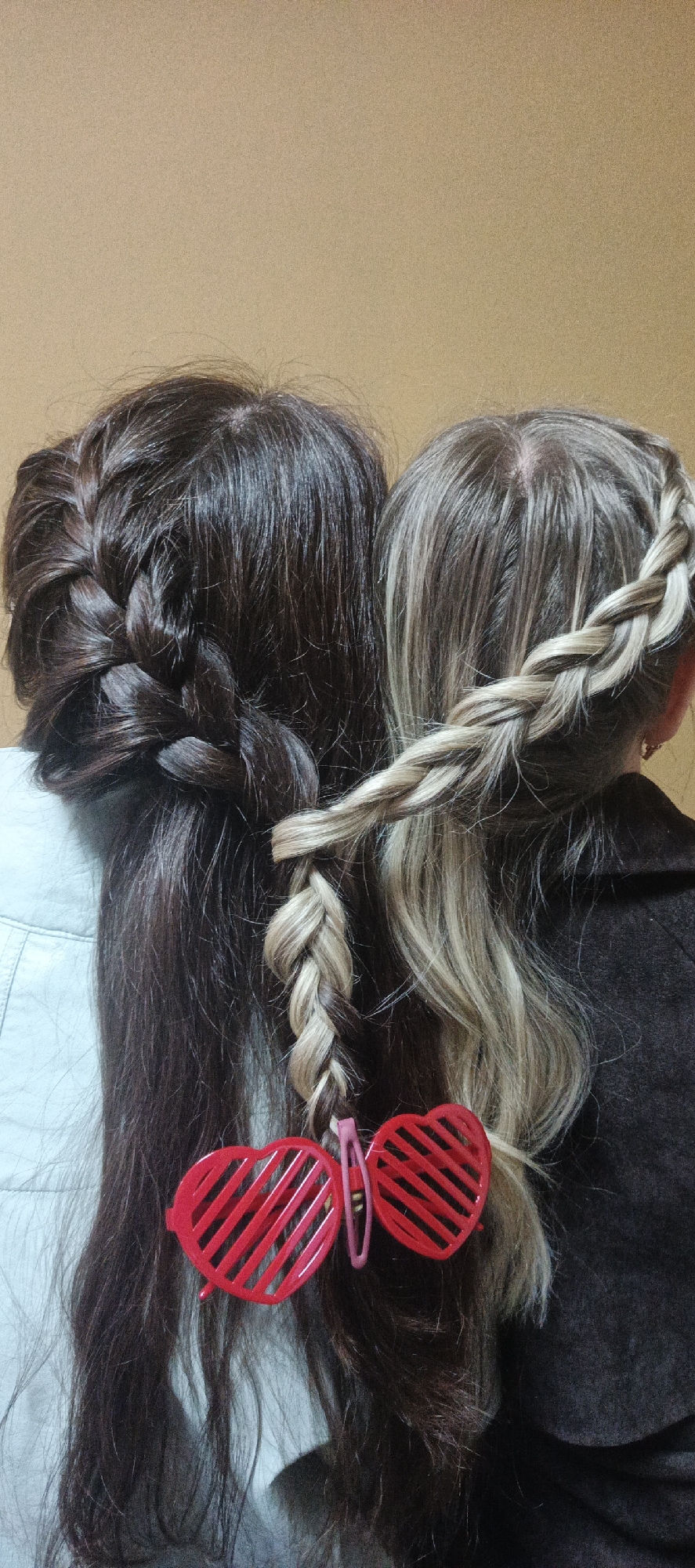 Two individuals, seen from behind, are the primary subjects, showcasing their distinct hairstyles. The person on the left has long, dark brown hair styled into an intricate, perhaps French or Dutch, braid that runs down the side of their head. They appear to be wearing a light-colored, possibly light blue or grey, garment.
The person on the right has long, highlighted hair, primarily light blonde and brown. Their hair features a prominent light-colored braid that originates from the side of their head. Notably, this lighter braid intertwines with a section of the dark-haired person's hair before continuing downwards. This shared element suggests a close connection or a styled "friendship" braid. The person on the right wears a dark, possibly brown, coat or top.
A pair of novelty red heart-shaped glasses, featuring horizontal slats, is clipped onto the intertwined sections of hair, held by a pink hair clip. The background is a plain, light brown wall, indicating an indoor setting. The image focuses on the creative hairstyling and the decorative glasses, evoking a sense of playfulness or connection. No text is visible in the image. The location context provided is Chaikovsky, Russia.