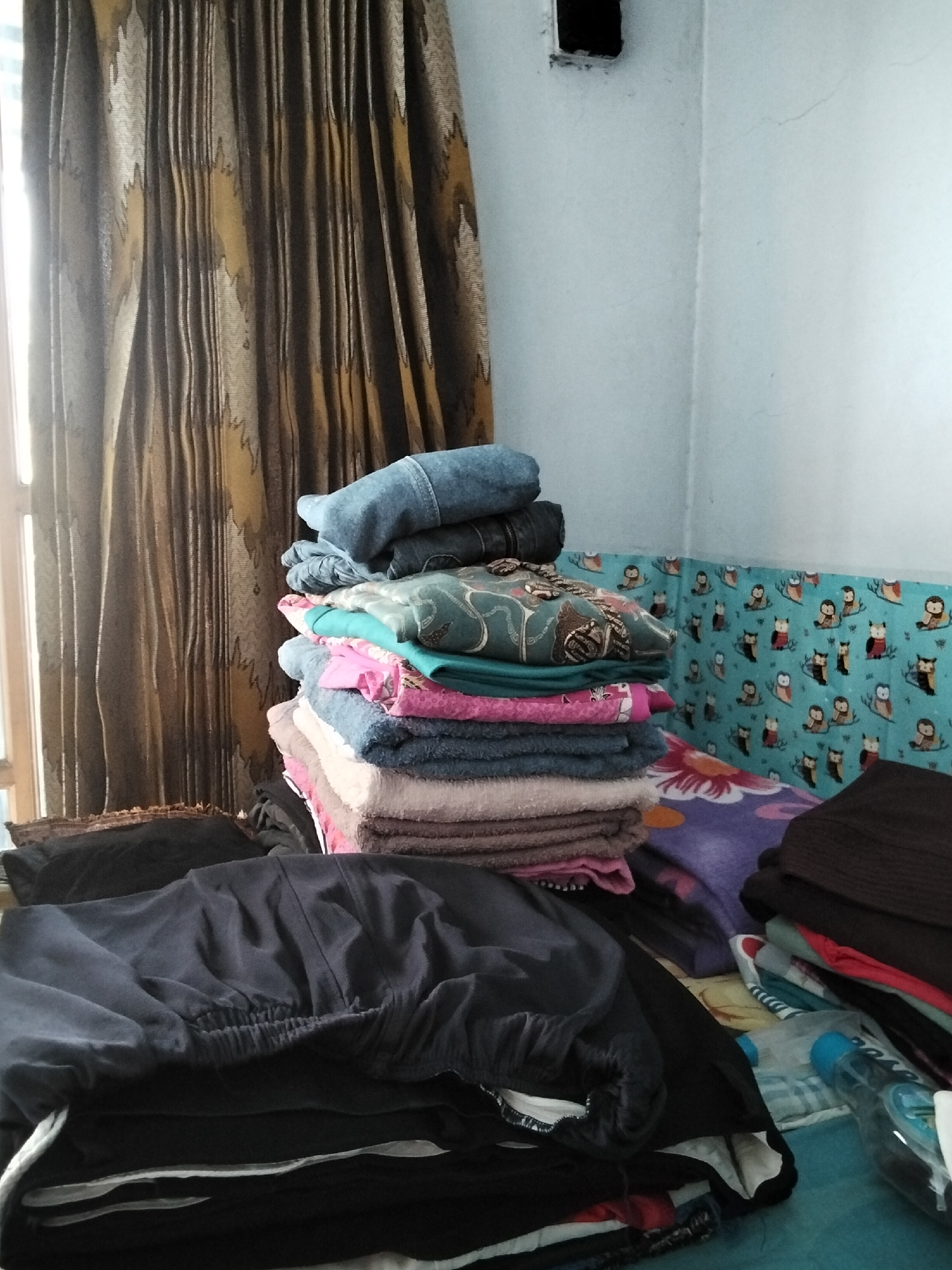 The image depicts an indoor scene, likely a bedroom, with a stack of folded clothes on a bed. The clothes include several folded towels in various shades of blue, pink, and beige, topped with a stack of folded denim jeans. To the left, a curtain with a patterned design of vertical stripes in shades of brown and yellow is visible, with a window allowing light to enter. In the foreground, a pile of dark-colored clothing, possibly pants or a skirt, is laid out. The background features a wall with a light blue patterned wallpaper adorned with cartoonish owl figures. A plastic bottle with a blue cap is also present among the folded items. The scene appears to be in a residential setting, and given the mention of Paguyangan, Indonesia, it is a domestic interior.