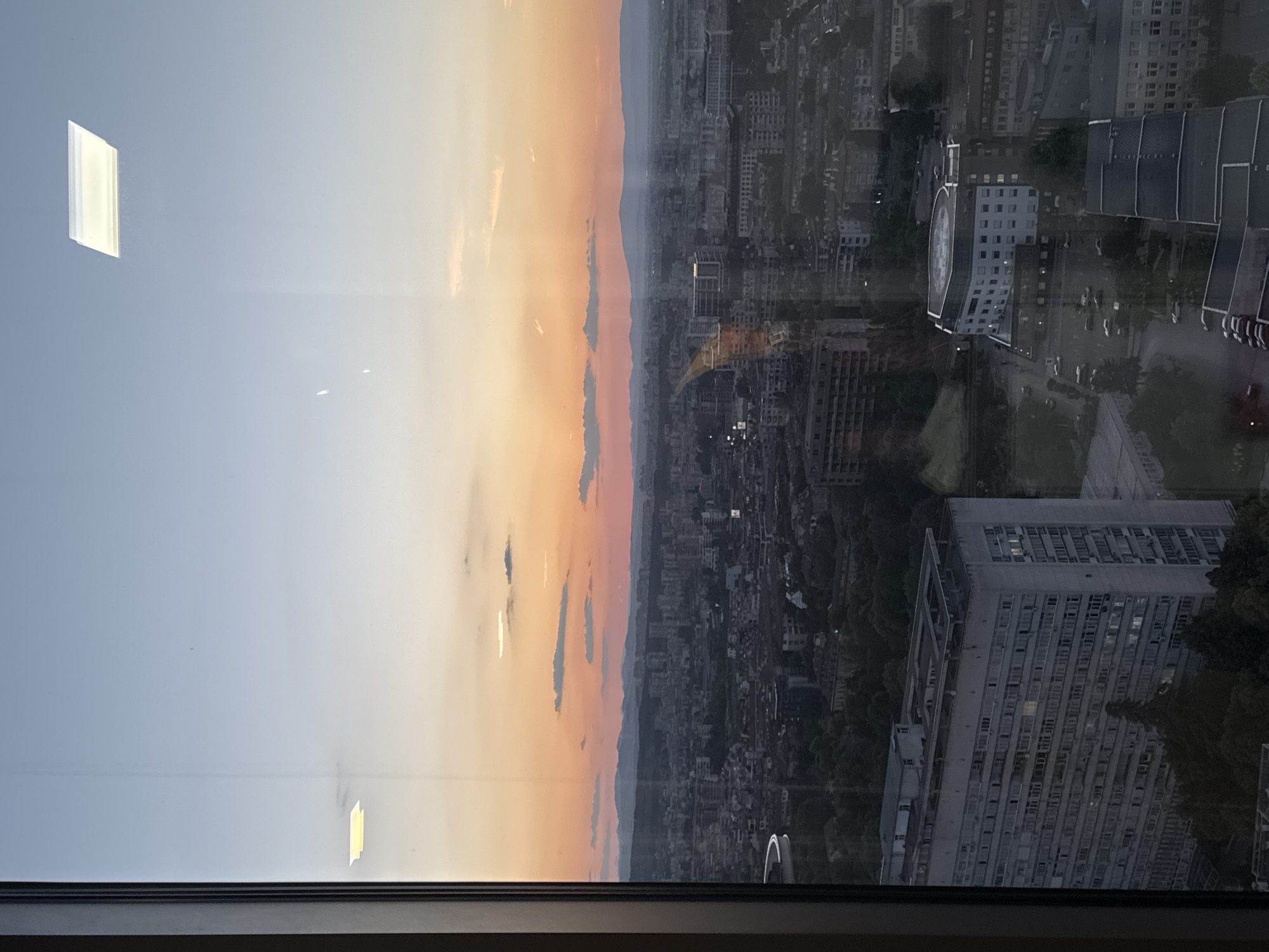 The image is an aerial view of Sofia, Bulgaria, captured during sunset. The sky displays a gradient of colors, transitioning from a pale blue at the top to warm oranges and pinks near the horizon, with a few scattered clouds. Below the sky, the city sprawls out with numerous buildings of varying heights and architectural styles. In the foreground, a large, light-colored, block-style building with many windows is prominent. Further into the city, other buildings and green spaces are visible. The scene is viewed through a window, evidenced by reflections and the window frame on the left side of the image. Two rectangular, illuminated light fixtures are visible as reflections on the window glass in the upper portion of the image. The overall impression is of an urban landscape at dusk.