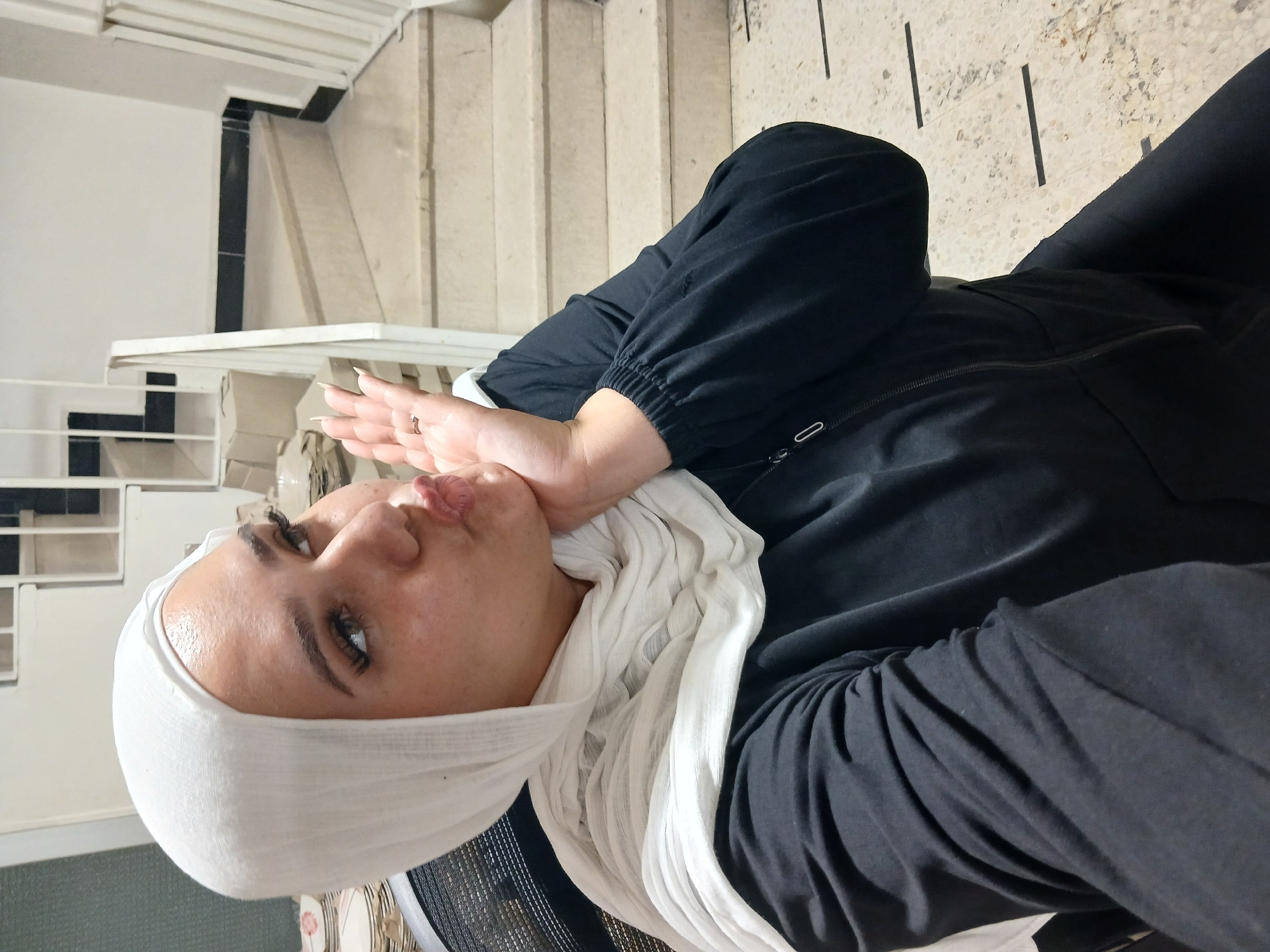 A medium shot photograph captures a young woman wearing a white hijab and a black zip-up jacket, seated in an office chair. Her right hand is raised to her chin, and she is puckering her lips as if to give a kiss. Her eyes are looking upwards and to the left, with dark eyeliner and long eyelashes. The background features a staircase with cream-colored steps and a white railing, possibly in a communal area within the Rukn ad-Din Municipality in Syria. Cardboard boxes are visible on a shelf to the left of the stairs. The floor is tiled with a speckled pattern and marked with black lines.