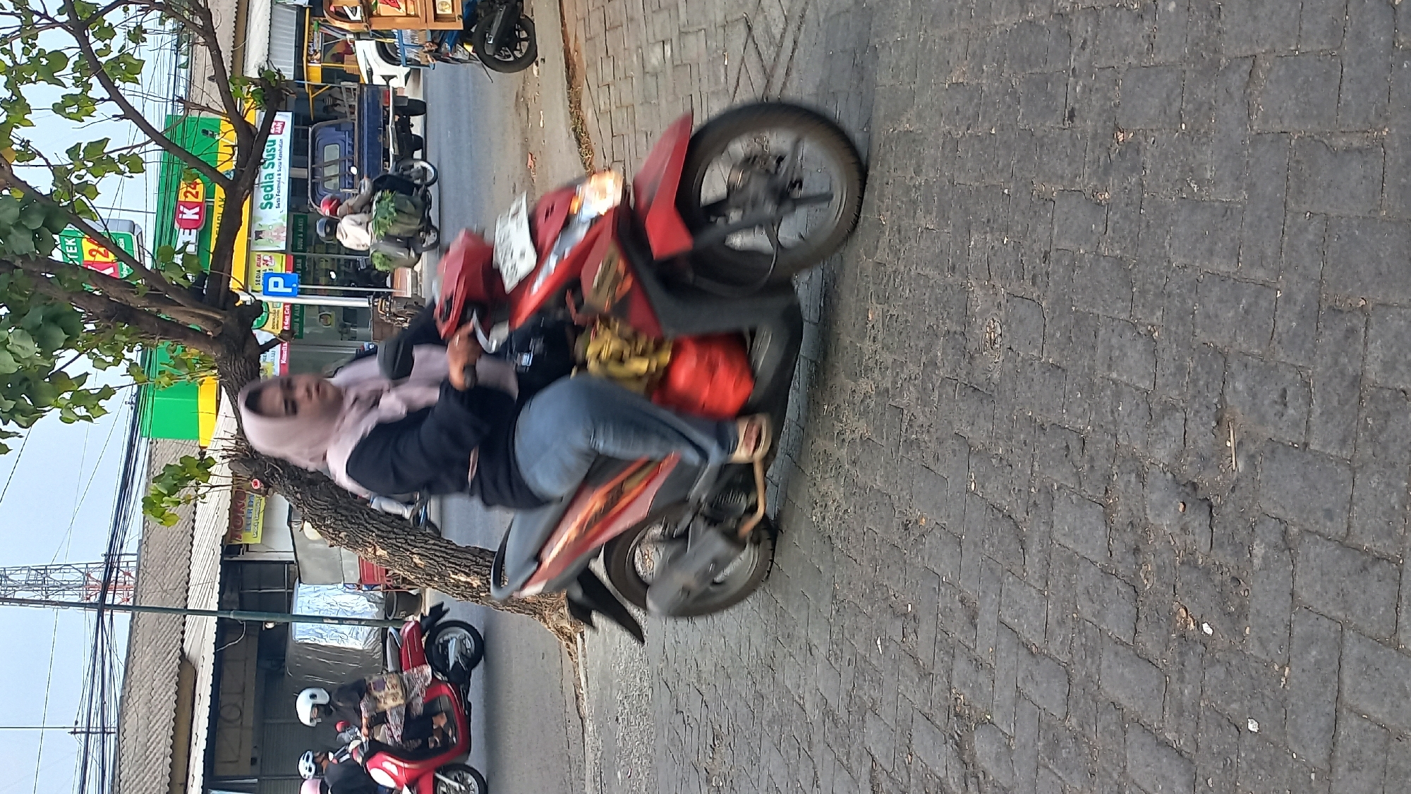 The image depicts a street scene in Mangunsari, Indonesia, featuring a woman riding a red scooter. The woman is wearing a light pink hijab and blue jeans, with her hands on the scooter's handlebars. The scooter is red with black accents and appears to be carrying a yellow bag on the back and a red jerrycan on the side. The scooter is moving, as indicated by the motion blur on the wheels and the rider. The street is paved with dark grey cobblestones. In the background, there are buildings with green and white signage, including one that reads "EK 24". Several other scooters and a blue pickup truck are visible in the distance, suggesting an urban environment. A tree with green leaves is positioned to the left of the central scooter. The sky is overcast.