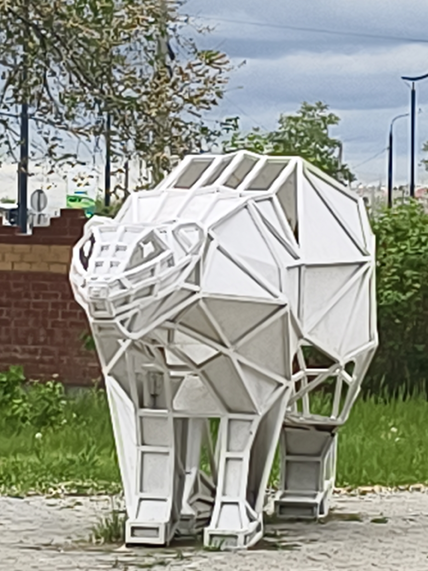 The image shows a geometric polar bear sculpture in Blagoveshchensk, Russia. The sculpture is made of white metal framing with filled panels and has a cubist, polygonal design. It stands on concrete paving with grass patches surrounding it. In the background, there's a brick wall, a road sign, trees, streetlights, and a cloudy sky.