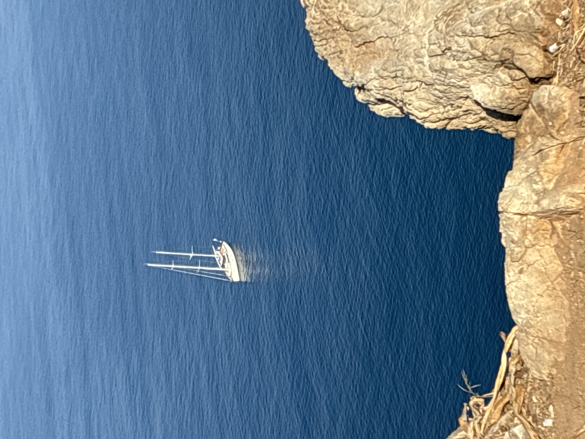An aerial, high-angle shot captures a white sailboat with two masts sailing on a calm, deep blue sea. The sailboat is positioned slightly right of center and appears to be a sloop with its sails down. In the foreground, the rough, textured surface of a tan-colored cliff face dominates the lower portion of the image, obscuring the shoreline. The cliff exhibits shadows and crevices, indicating an uneven terrain. The overall scene suggests a tranquil nautical setting near a coastal area. This scene is located in Alanya, Turkey, a city renowned for its stunning coastline and historical cliffs.
