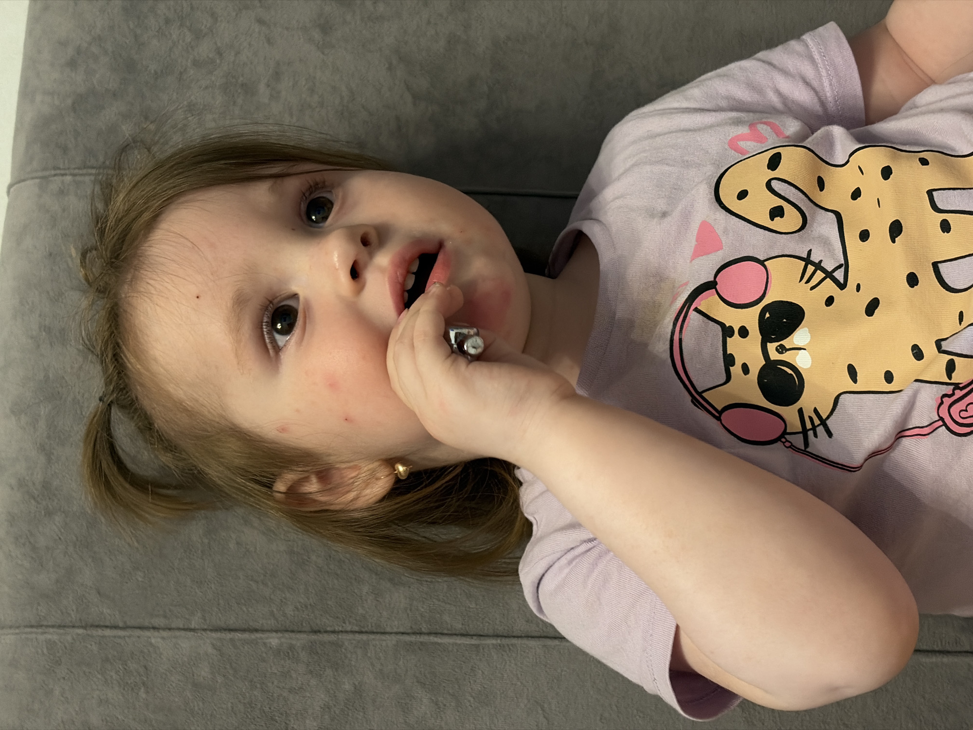A young girl with light brown hair is being photographed indoors, likely in a residential setting in Chaikovsky, Russia. She is wearing a light purple t-shirt with a cartoon cheetah wearing headphones and sunglasses printed on the front. The girl has her right hand near her mouth, holding a small, silver cylindrical object, possibly a crayon or lipstick. Her eyes are wide open and looking slightly to the right. The background is a textured gray surface, likely upholstery or a wall. There are red marks on her cheeks and around her mouth.