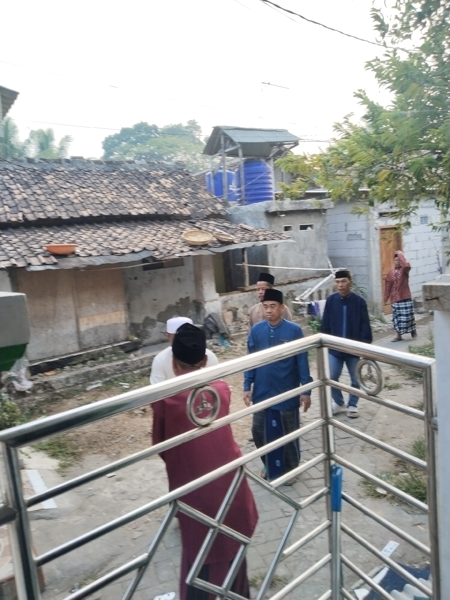 A group of individuals, appearing to be mostly male and some wearing traditional religious attire, are gathered outdoors in what seems to be a residential area. The environment includes buildings with tiled roofs, a visible water tank, and a metal railing in the foreground. The context of Serang, Indonesia, suggests a possibly warm climate and a cultural setting where such attire is common. The group's focus is unclear, but they are positioned in a walkway or courtyard.
