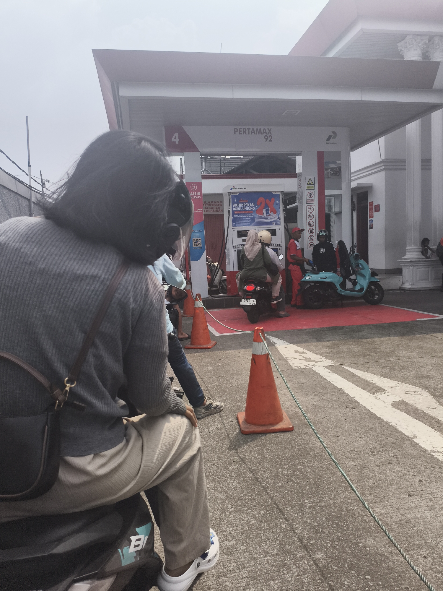 The image captures a scene at a Pertamina gas station, likely in the city of Depok, Indonesia, indicated by a gas pump displaying "PERTAMAX 92" and advertising in Indonesian. In the foreground, a person is seated on a motorcycle, facing away from the camera. This individual wears a grey knitted sweater, light-colored pants, and white clogs. A dark brown crossbody bag is slung over their shoulder.
Behind this person, at a gas pump labeled with the number '4', a Pertamina attendant in a red uniform is interacting with two individuals on another motorcycle. The attendant is speaking to a person wearing a black helmet and a black jacket, who is seated behind the driver of the motorcycle. The driver of this motorcycle wears a light blue shirt and is partially obscured by the attendant.
To the right of the gas pump, a light blue scooter is parked, with a person in a red uniform standing nearby. Several orange traffic cones are placed on the ground, one in the mid-ground and another in the foreground, with a rope extending between them. The gas station canopy is visible overhead, supported by white pillars. The ground is asphalt with white painted lines. The sky appears overcast.