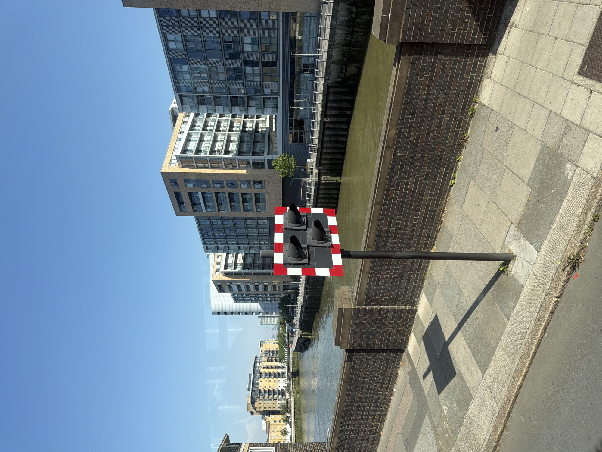 The image shows a canal in London, United Kingdom, with buildings on either side. A traffic signal, black with a red and white striped border, stands on the edge of the canal. The water in the canal is murky. Buildings vary in height and style. The sky is clear and blue. The viewpoint appears to be from slightly above street level, possibly from inside a vehicle. The scene is well-lit with sunlight, casting shadows from the traffic signal.