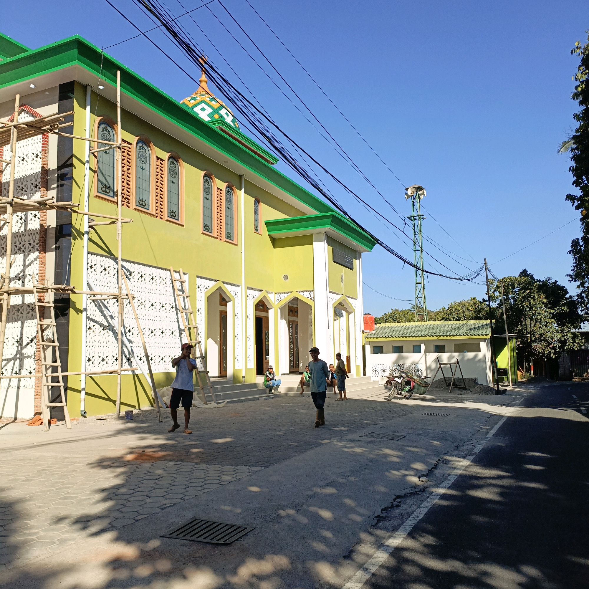 The image depicts the exterior of a mosque in Salatiga, Indonesia, possibly under construction or renovation, given the presence of scaffolding and bamboo poles. The building is predominantly lime green with white accents around the entrances and decorative white latticework on the exterior walls. The mosque features arched doorways and windows with stained glass. A vibrant, multi-colored dome, green and white with orange accents, crowns a section of the building.
In the foreground, several individuals are present. Two men are walking on the paved area in front of the mosque, one in a blue t-shirt and shorts, the other in a grey t-shirt and dark pants. Other people are seen sitting on the stairs leading up to the mosque's entrances. To the right, a motorcycle is parked near a smaller building that also has a green roof and white latticework.
The environment appears to be a public street, with asphalt pavement visible in the lower right portion of the image. A green transmission tower with speakers is visible in the background, along with trees and a clear blue sky. Overhead, a complex network of black electrical wires crisscrosses the sky. The lighting suggests it is daytime, with shadows cast on the ground by the building and overhead wires.