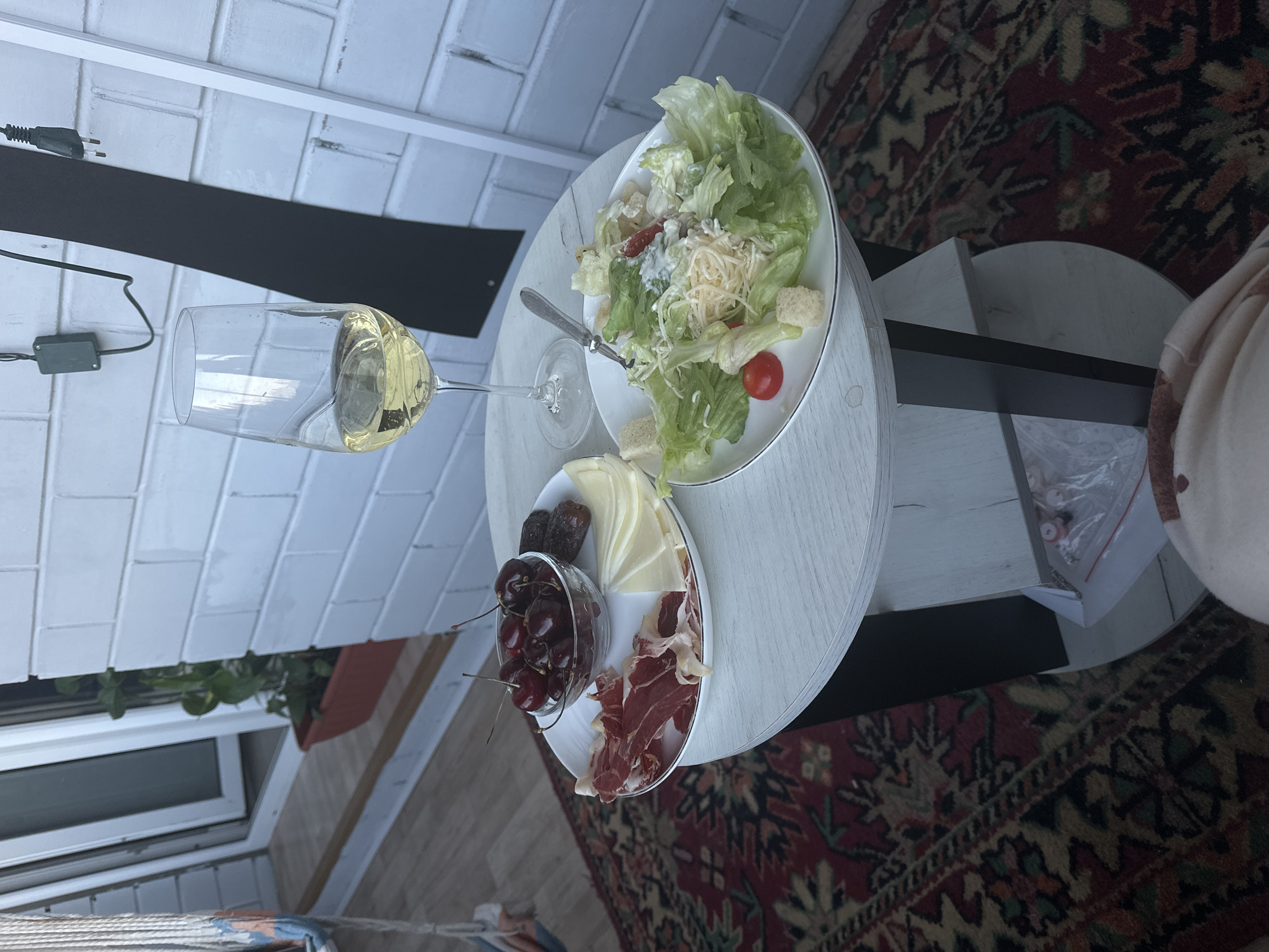 An angled overhead shot captures a small, round, white table set with a meal, likely on an outdoor patio or balcony. A tall glass of white wine sits alongside a white plate holding a Caesar salad composed of lettuce, croutons, cherry tomatoes, and shredded cheese. Another white plate serves as a charcuterie board, featuring slices of cured ham, wedges of yellow cheese, dried dates, and a glass bowl filled with fresh red cherries.
The table is positioned on a richly patterned rug with dominant red, green, and beige hues. In the background, a white painted brick wall is visible. An electrical cord with a two-pronged plug and a small, green control box extends from the left. Further back, a window is partially seen, with a planter box containing green foliage above it. To the far right, a light-colored blanket or throw is casually draped over an unseen object. A clear plastic bag containing small white items, with the Russian word "РАССМОТРИТЕ" (meaning "CONSIDER" or "EXAMINE") printed on it, is on the floor near the table. The scene suggests a relaxed daytime meal or snack.