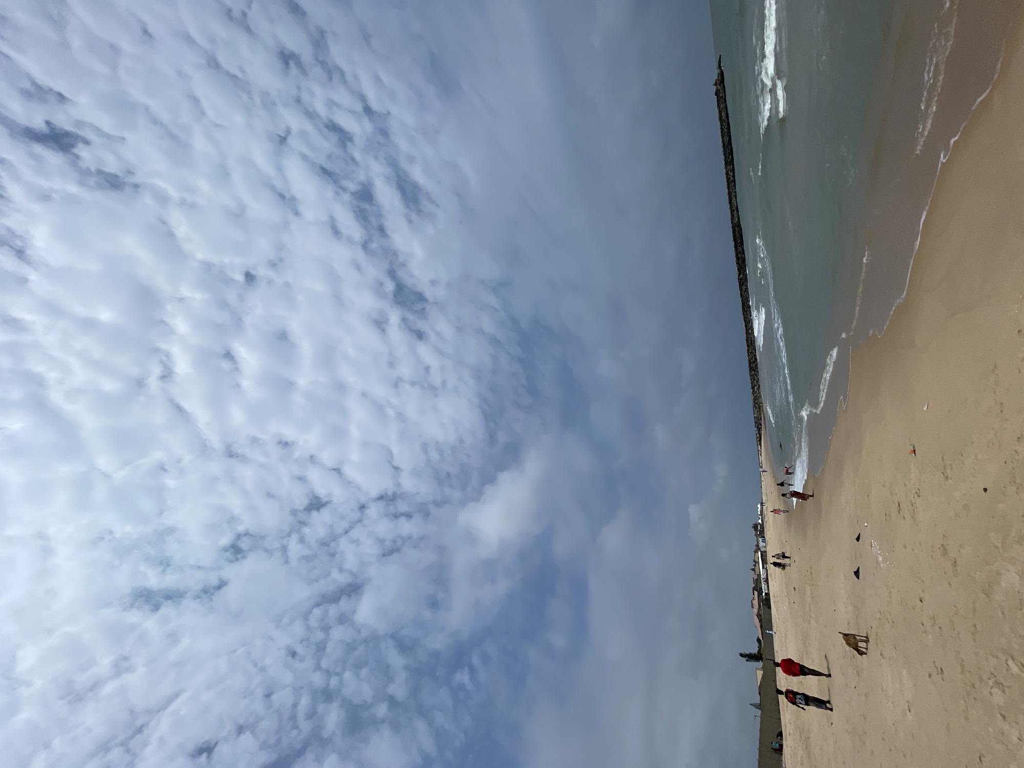 The image depicts a beach scene in Ilado, Nigeria, under a cloudy sky. The foreground is a sandy beach with footprints and some scattered debris, including a plastic bottle. Several people are present on the beach: two men are walking towards the left, one in a red shirt and the other in a dark shirt with the number 23. Further down the beach, more people are walking or standing, and a dog is visible in the sand. In the background, there are some buildings and structures, possibly houses or small shops. The sea is to the right, with waves rolling onto the shore. A long, man-made stone breakwater extends into the sea from the right side of the image, with waves breaking against it. The overall atmosphere suggests a slightly overcast day at the coast.