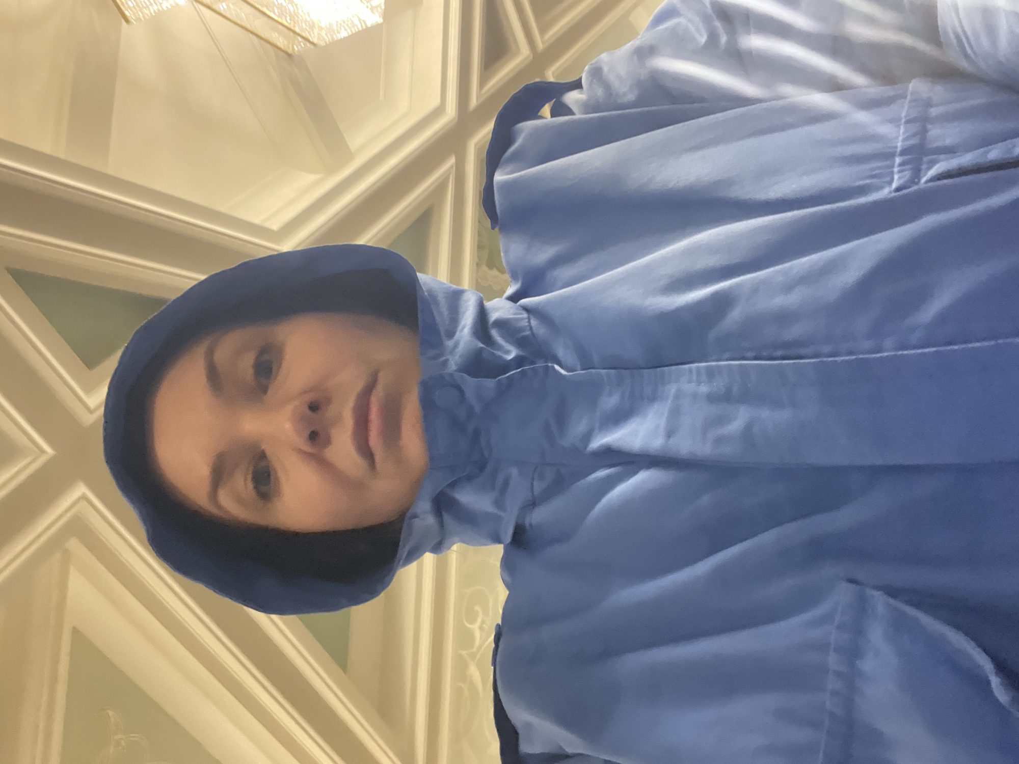 A selfie, possibly taken by a woman, shows her face framed by a blue hooded jacket. She has light skin and appears to be indoors. Behind her, the background includes a section of the ceiling and upper walls, which feature ornate white trim and a pale yellow or beige color. A chandelier is visible at the top of the frame. The setting seems to be an interior space with classical architectural details. Given the mention of Kazan, Russia, it is possible this is within a building known for such decoration in the city.