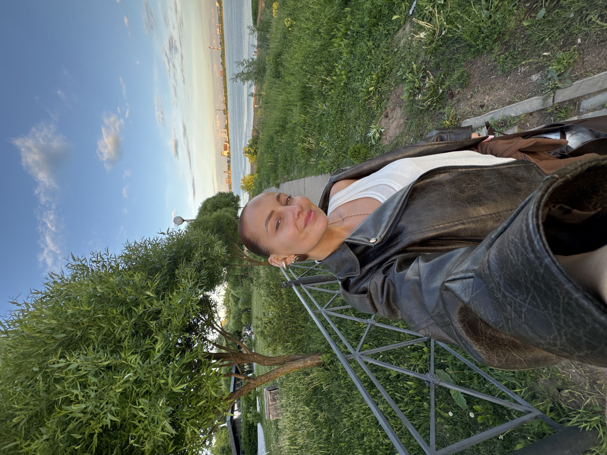 This image captures a young woman in a relaxed pose outdoors, likely taking a selfie, in a park-like setting alongside a large body of water in Izhevsk, Russia. The woman, with her hair pulled back, is looking directly at the camera with a pleasant, soft smile. She is dressed in a white tank top under a dark, possibly black or very dark brown, leather jacket. Multiple earrings adorn her left ear, and a delicate, beaded necklace is visible.
She appears to be seated or leaning on a dark metal structure, possibly part of a bench or railing, which occupies the lower right portion of the frame. The foreground features lush green grass and a dirt path. To the left, a vibrant green tree with weeping willow-like foliage frames the scene.
In the mid-ground, a wide body of water extends across the frame. On the distant horizon, across the water, a cityscape with various buildings and industrial elements is visible. The sky is a clear, light blue with scattered, wispy white clouds. The lighting suggests late afternoon or early evening, casting a soft, warm glow over the landscape, indicative of fair weather. The overall mood conveyed is calm and content. No visible text is present in the image.