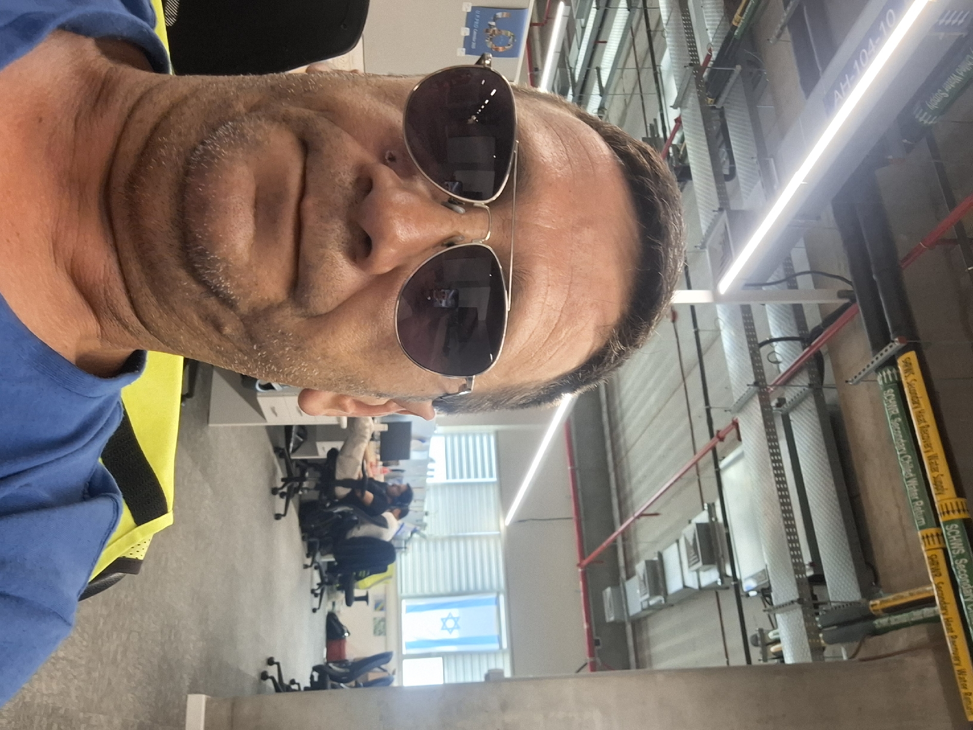 The image shows a man in the foreground, wearing sunglasses and a blue shirt over a yellow high-visibility vest. He is looking directly at the camera. In the background, an office environment is visible with cubicles, desks, chairs, and computers. An Israeli flag is hanging in a window. The ceiling features exposed pipes and lighting fixtures. The overall impression is of a man taking a selfie in an office setting. The image is taken in the city of Kiryat Gat, Israel.