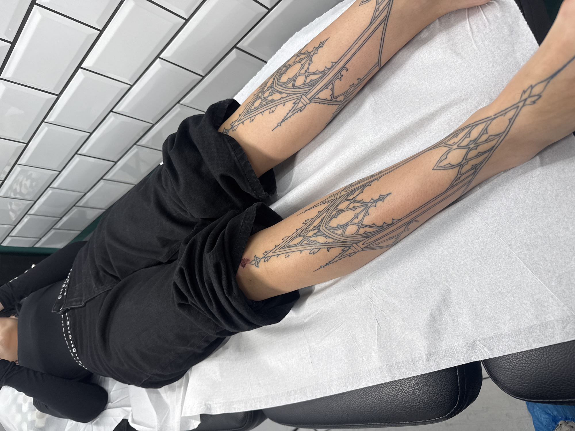 A person is lying on their back on a black upholstered bed or chair, covered with a white disposable paper sheet, in what appears to be a tattoo studio located in Sofia, Bulgaria. The individual is wearing black casual clothing, including jeans and a long-sleeved top, with their arms extended upwards.
The primary focus is on the intricate line-art tattoos on both forearms. These tattoos feature an elongated, symmetrical design with geometric and gothic-inspired patterns, reminiscent of architectural tracery or ornate window frames. The detailed black lines cover a significant portion of the inner forearms, extending from the wrists towards the elbows. A small, reddish mark or smudge is visible on the inner left forearm, near one of the tattoo lines, indicating a fresh tattoo or minor irritation.
The background features a wall covered with glossy white rectangular subway tiles set with dark grout, suggesting a clean and modern environment. The overall lighting is bright and uniform, characteristic of a professional studio. A blue shoe cover is partially visible on the floor at the bottom right of the image. No text is visible.