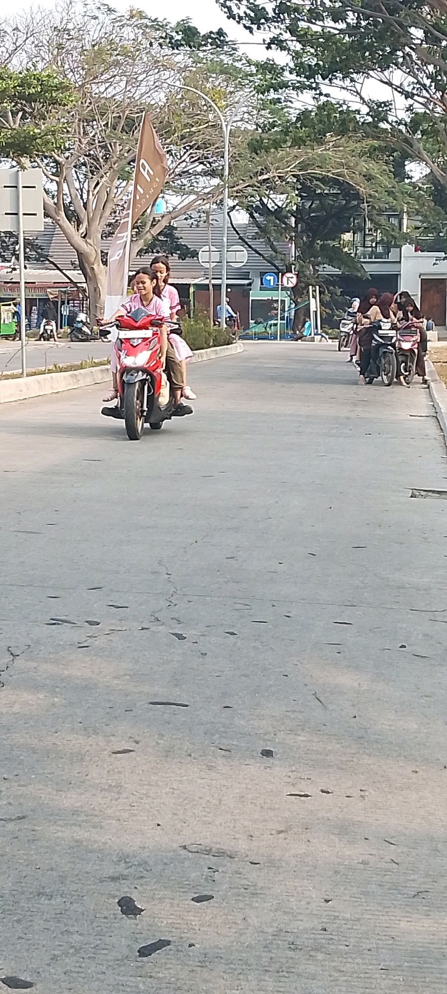 A front-facing medium shot captures two young Indonesian women riding a red motorcycle on a paved road in Satriamekar, Indonesia. The woman in front, wearing a pink t-shirt and brown pants, is smiling and looking forward. The woman behind her, also in a pink t-shirt and skirt, leans forward slightly. They are in motion, with the motorcycle's front wheel turned to the left. The road is gray and cracked, with some debris. In the background, trees with sparse green leaves line the street. Several other motorcycles with multiple passengers are visible further down the road, suggesting a bustling street. Buildings with white and gray facades are partially obscured by the trees. A brown flag with white lettering, possibly a company logo, is planted on the left side of the road. The lighting suggests it is daytime, with the sun casting shadows.