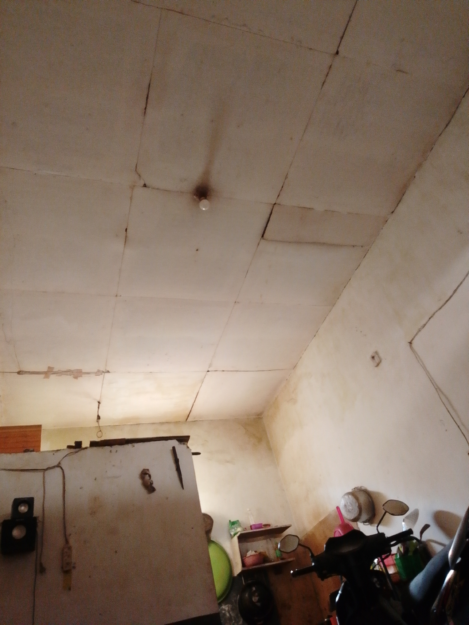 The image shows a dimly lit, unfinished interior space, possibly a storage area or workshop. The ceiling is constructed of off-white panels with visible cracks and signs of wear. A bare light bulb hangs from the ceiling in the upper central portion of the frame. The walls are also off-white and appear stained and discolored in places.
On the left side of the image, a large, off-white cabinet or refrigerator dominates the scene. A black speaker is mounted on its left side. Various wires and a power outlet are visible on the wall behind the cabinet. A light brown wooden panel is mounted on the wall above the cabinet.
The right side of the image features a parked motorcycle, partially visible, with its handlebars, mirrors, and front portion in focus. Behind the motorcycle, there are shelves with various items, including containers and what appears to be a pink watering can. The floor is not clearly visible due to the angle and lighting. The overall impression is of a cluttered and utilitarian space. The image is assumed to be located in Bohar, Indonesia, based on the provided context.