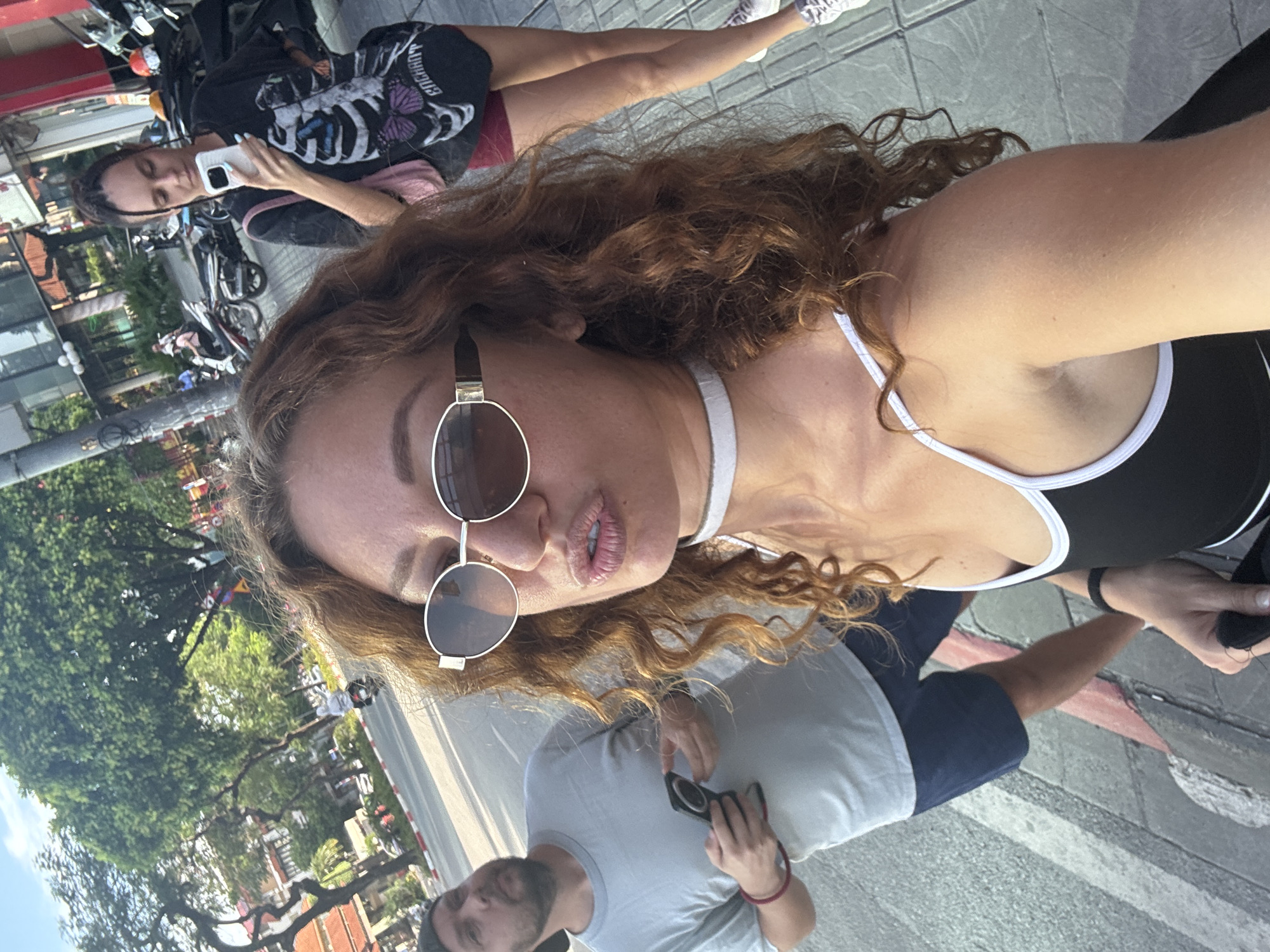 In an outdoor urban setting, likely in Hanoi, Vietnam, three individuals are captured in a photograph. The foreground is dominated by a woman taking a selfie, her face close to the camera with lips pursed. She has long, wavy auburn hair, is wearing large oval sunglasses with a gold frame, and a white choker necklace. Her attire includes a black and white-trimmed top. Behind her, a man wearing a grey t-shirt and dark shorts is partially visible, holding a dark object, possibly a phone, in his left hand. Further in the background, another woman with dark hair, styled in braids, is holding a white smartphone, seemingly also taking a photo. She is wearing a black t-shirt with a graphic print of a skeleton and butterflies, and dark red shorts. The environment suggests a street scene with parked motorcycles and trees lining a sidewalk. The overall lighting indicates it is daytime, with sunlight casting shadows.