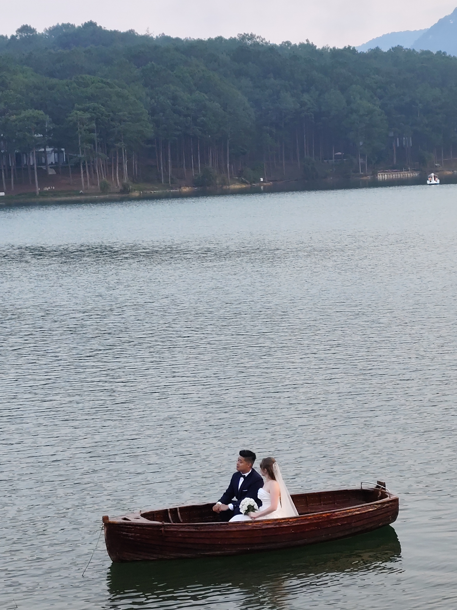 A low-angle, eye-level, full outdoor shot depicts a bride and groom in a wooden rowboat on Xuan Huong Lake in Da Lat, Vietnam. The couple is centered in the bottom of the frame. The groom, to the left, wears a navy suit and bow tie. The bride, to the right, wears a white wedding dress and a veil. She holds a bouquet of white flowers. The lake occupies the majority of the frame. In the background, a line of tall pine trees edges the shoreline. Several buildings are visible among the trees. To the right, a small white boat with people is docked at a wooden pier. The sky is overcast.