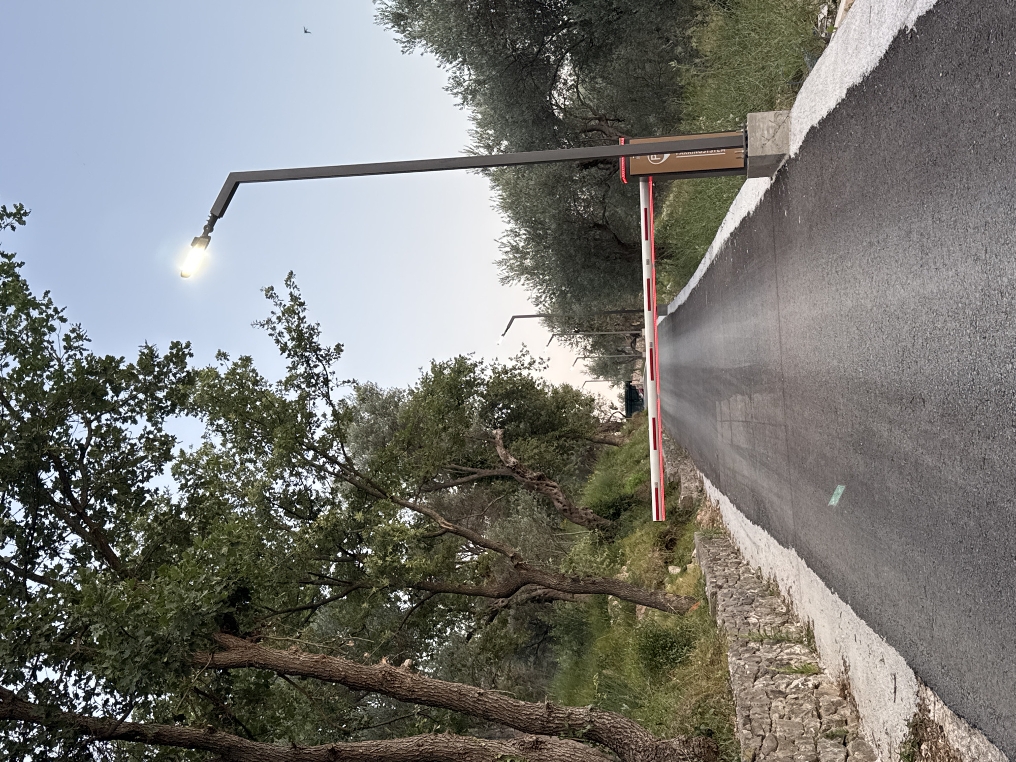 The image shows a paved road in Burtaiši, Montenegro, leading towards an automated barrier gate. The road is bordered by a stone wall on the left and lush green vegetation and trees on both sides. A street lamp with its light on is visible on the right side of the road, and several other street lamps are spaced along the road in the distance. The barrier gate is white with red reflective stripes and appears to be lowered, blocking the road. A sign with a circular emblem and text is attached to the post of the barrier gate. The sky is clear and a light blue, suggesting it is either early morning or late afternoon. There are no people visible in the image.