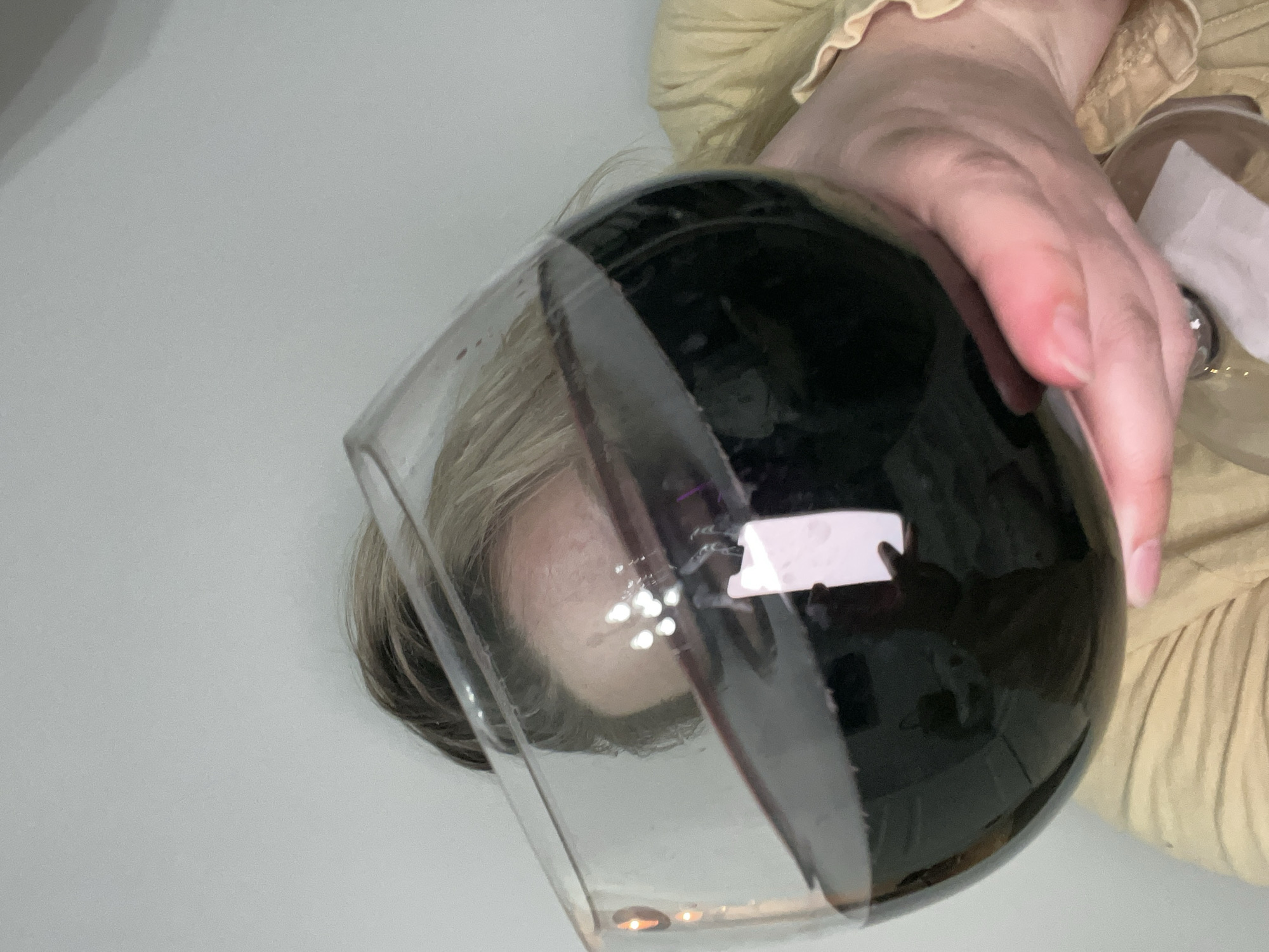 A person is holding a glass of dark liquid, likely wine. The person's face is visible through the glass. The person is wearing a tan-colored shirt with frills on the sleeve. Another glass or container is partially visible in the person's hand. The background is a plain white surface. The location cannot be determined from the provided image.