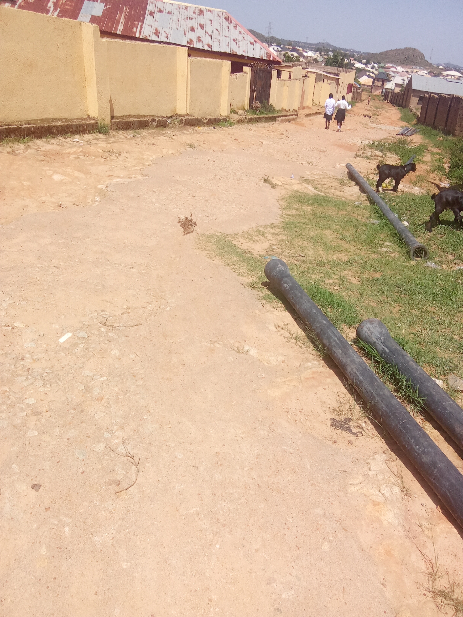 The image depicts a dusty, unpaved road in what appears to be a residential area, likely in Barakin Akawo, Nigeria. On the left side of the road, there is a long, light yellow wall with a corrugated metal roof visible above it. The wall is somewhat weathered and shows signs of age. The road surface is uneven and composed of dirt and gravel.
Further down the road, two individuals, appearing to be young males wearing white shirts and dark skirts or kilts, are walking away from the camera. To the right of the road, several black, cylindrical objects, possibly pipes or culverts, are laid on the ground, some partially covered by grass. Two black goats are also present in this area, with one actively grazing. In the background, a cluster of buildings with varied roofing can be seen, extending up a gentle incline, suggesting a settlement in a hilly or elevated terrain. The sky is clear and blue, indicating a sunny day. The overall scene suggests a somewhat unkempt, possibly developing, urban or semi-urban environment.