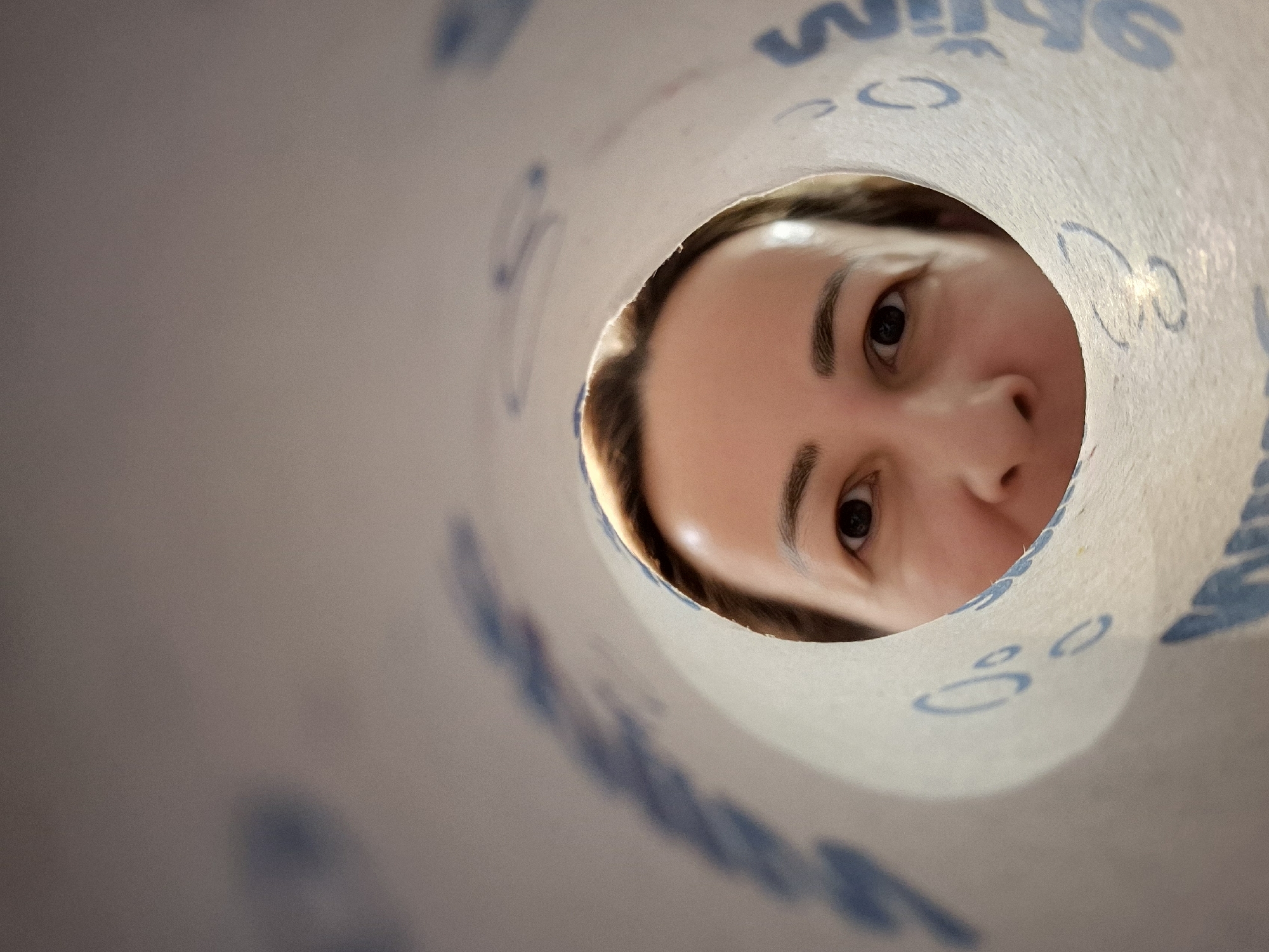 A woman's face is visible through the opening of a white roll of paper that has blue text and symbols printed on it. The woman is looking directly at the camera. Her left eye is in focus, and her right eye is partially visible. The paper roll is in the foreground, and the woman's face is in the background. The lighting is soft, and the colors are muted.