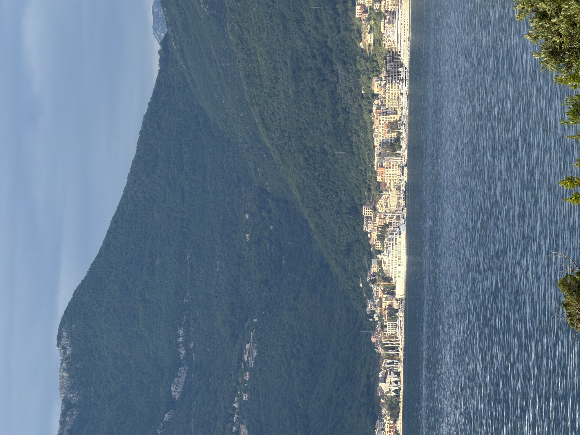 The image shows a view of the city of Krašići, Montenegro, located along the coast of the Adriatic Sea. A tall, green mountain slopes down towards the coastline, where a dense cluster of buildings, including residential and commercial structures, runs parallel to the water's edge. The sea is a dark blue, with subtle ripples creating a textured surface. A tree branch with green foliage is visible in the upper right corner of the image. The sky is a light blue with faint cloud formations in the upper left. The image appears to be taken from an elevated vantage point.