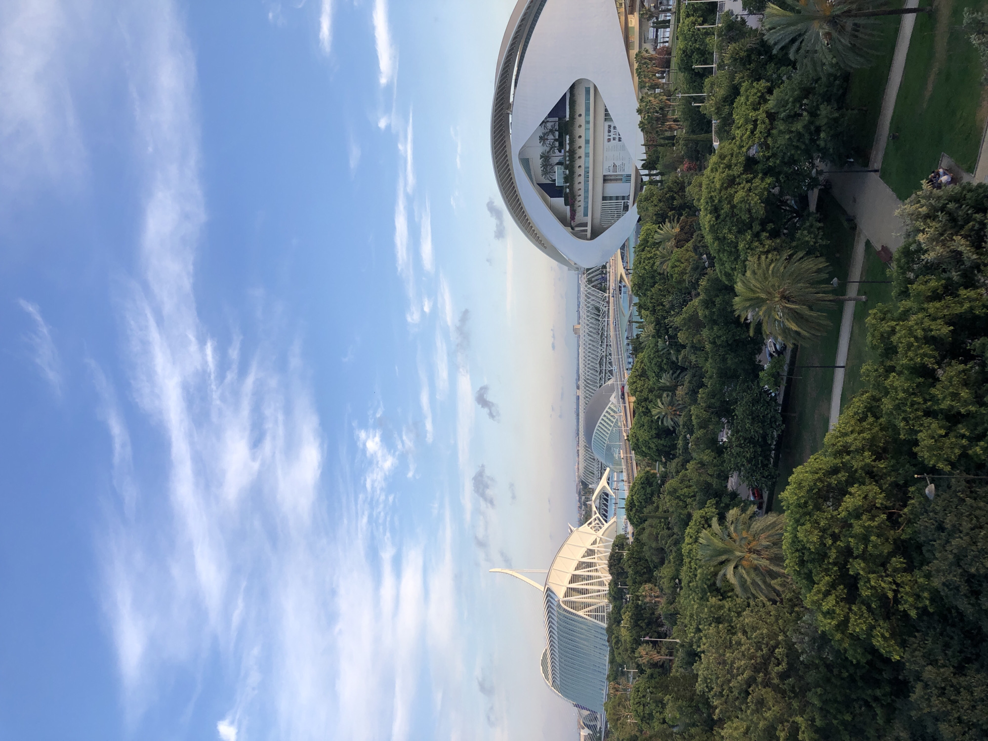 This aerial, high-angle, wide shot captures the City of Arts and Sciences in Valencia, Spain, bathed in the soft light of late afternoon or early evening. The sky is a gradient of blue, transitioning from a bright azure overhead to a paler hue near the horizon, dotted with wispy white clouds.
Dominating the midground are two of the complex's iconic structures: the Hemisfèric, a bold, eye-shaped building with a white, curved exterior, and the Palau de les Arts Reina Sofía, characterized by its large, white, boat-like canopy. The Hemisfèric's façade displays a large advertisement for an IMAX experience. In the distance, the L'Oceanogràfic, another part of the complex, is visible, its white, arched structures blending into the cityscape. These architectural marvels are surrounded by a lush, verdant expanse of trees, including palm trees, creating a striking contrast between nature and modern architecture. Pathways and grassy areas are interspersed within the greenery, with a few indistinct figures visible in the lower right portion of the image, appearing to be seated on a bench. Cars can be seen on elevated roadways that weave through the park-like setting. The overall scene conveys a sense of spaciousness and modern design integrated harmoniously with nature.